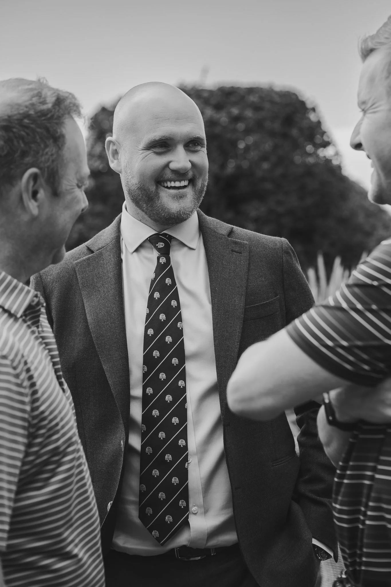 Four men standing outdoors, engaged in conversation. The man in the center is bald, smiling, and dressed in a suit with a patterned tie. The other men are partially visible, wearing casual shirts, and are smiling or laughing.