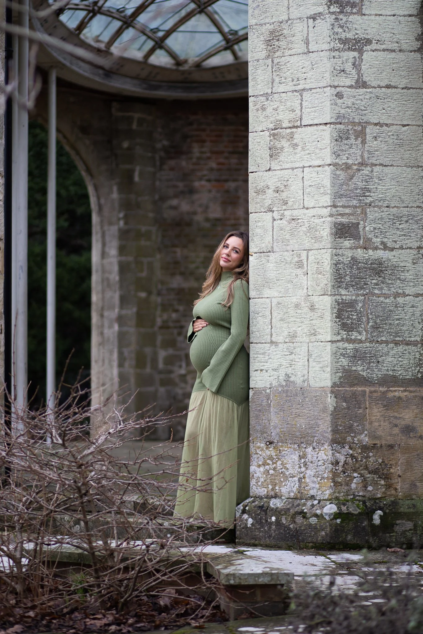 Pregnant woman standing outdoors behind a brick and stone structure, wearing a green dress.