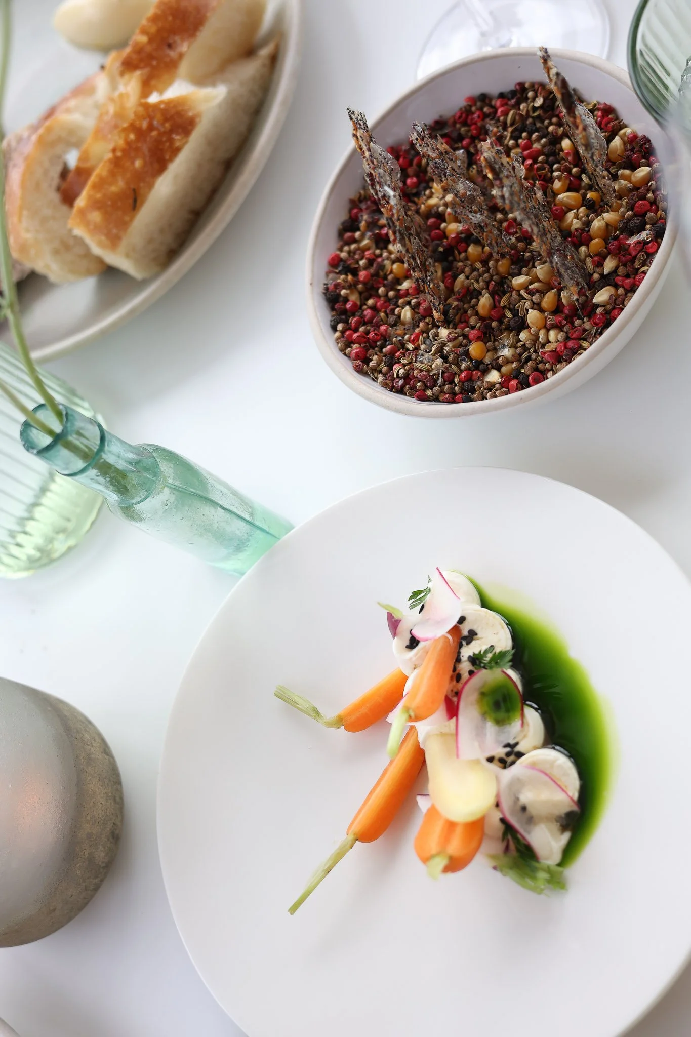 A plate with colorful vegetable and seafood dish garnished with herbs, served with carrot sticks and radish slices. An abstract green sauce surrounds the seafood.