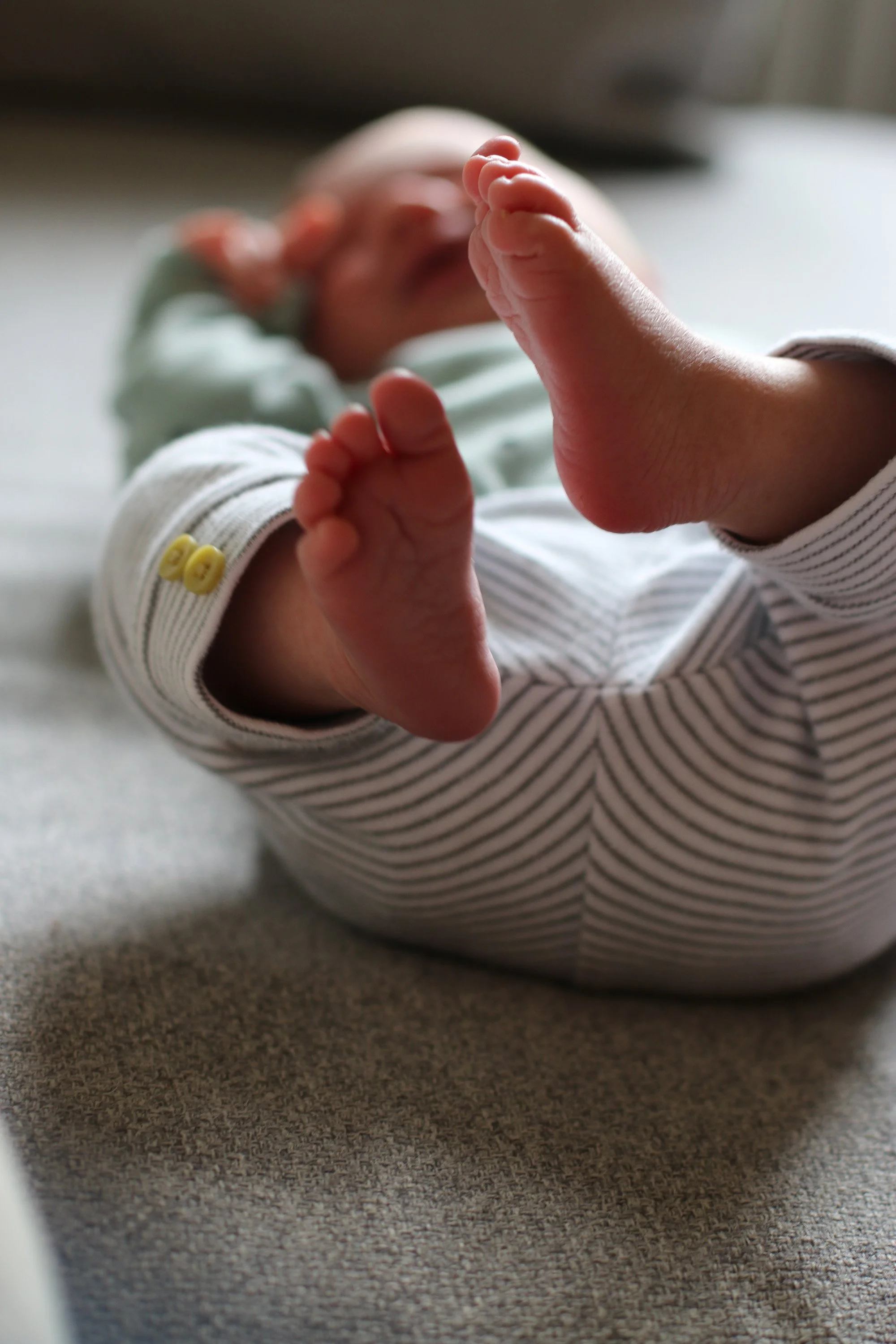 Close-up of a baby's hand with tiny fingers, reaching out towards the camera, in a soft focus with a blurred face. The baby is wearing a striped long-sleeve outfit with yellow buttons.