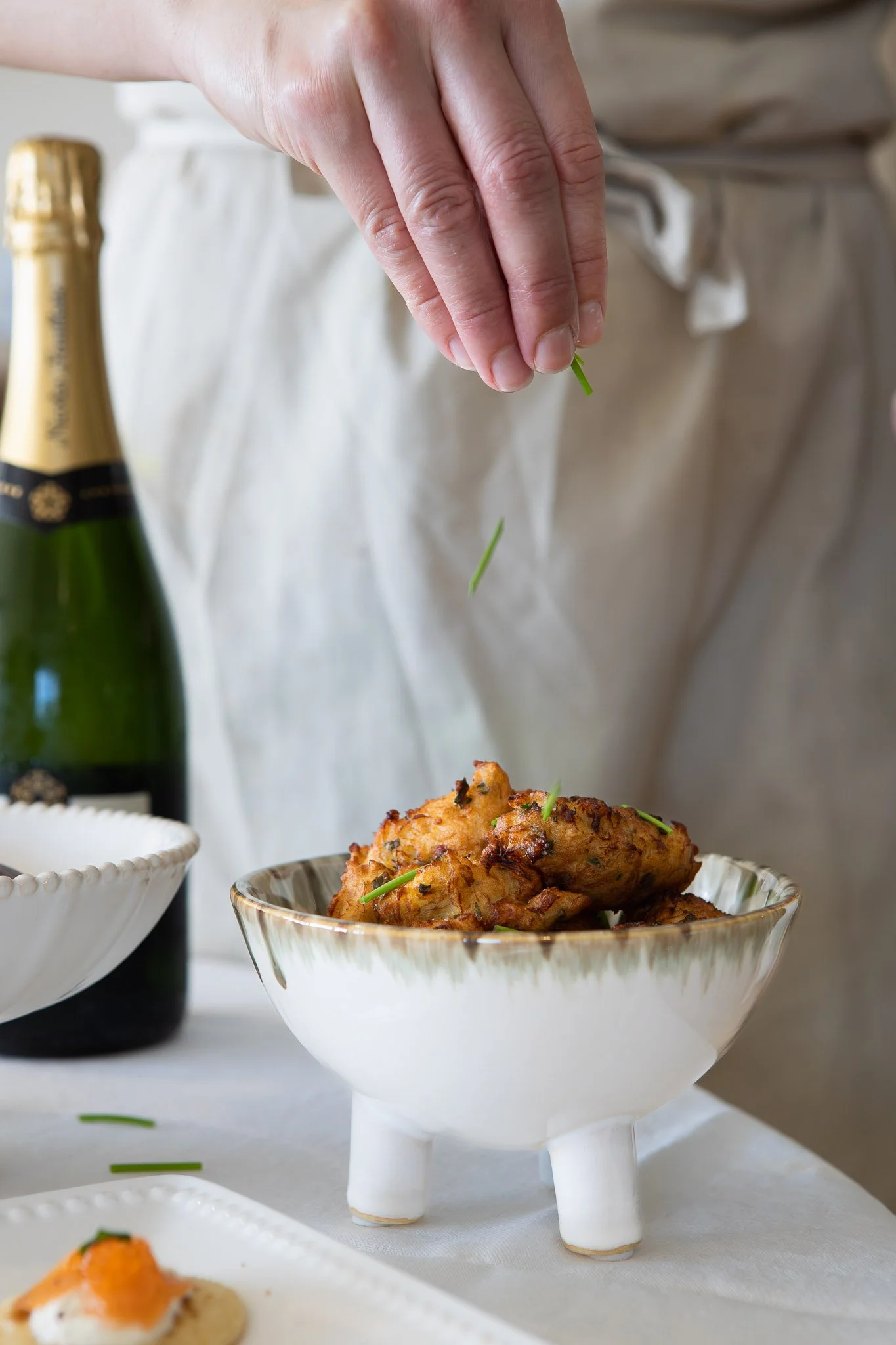 Person placing a green garnish on a plate of fried chicken, with a champagne bottle and empty bowls on a white tablecloth.