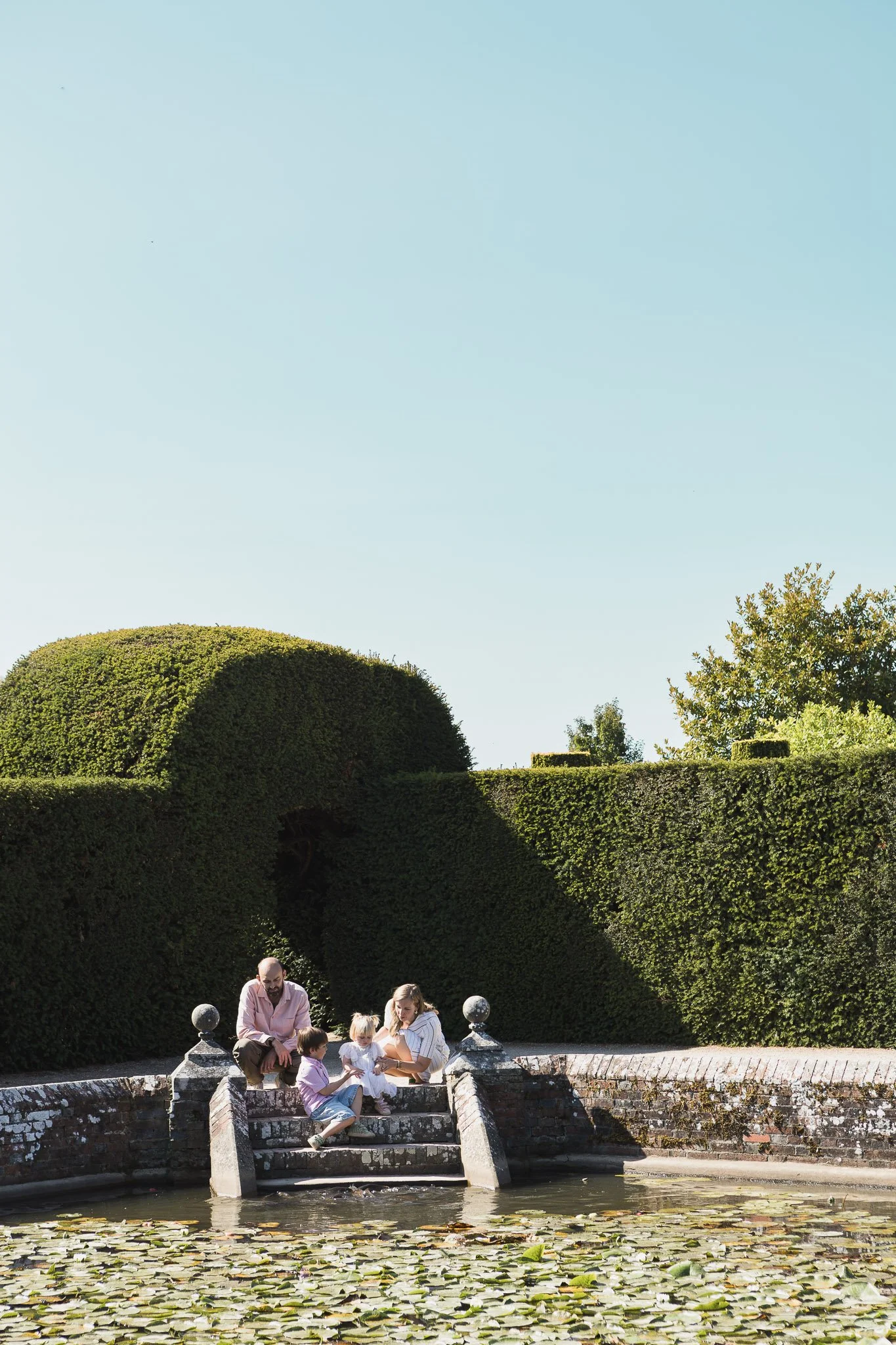 Family sitting on a stone staircase by a pond with water lilies, surrounded by lush green topiary and trees under a clear blue sky.