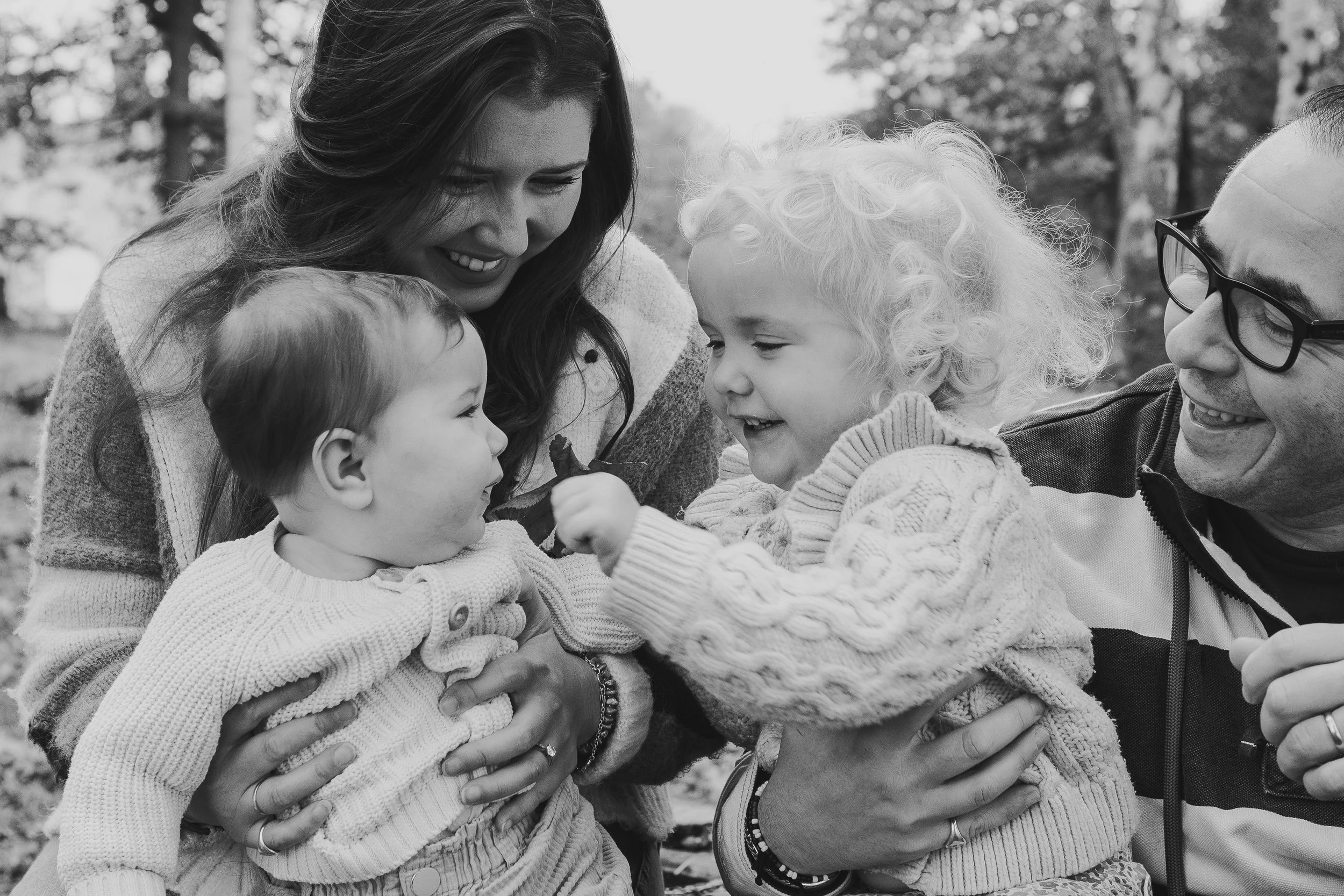 A black-and-white photo of a family of four smiling and hugging outdoors, with a blurred background of trees.