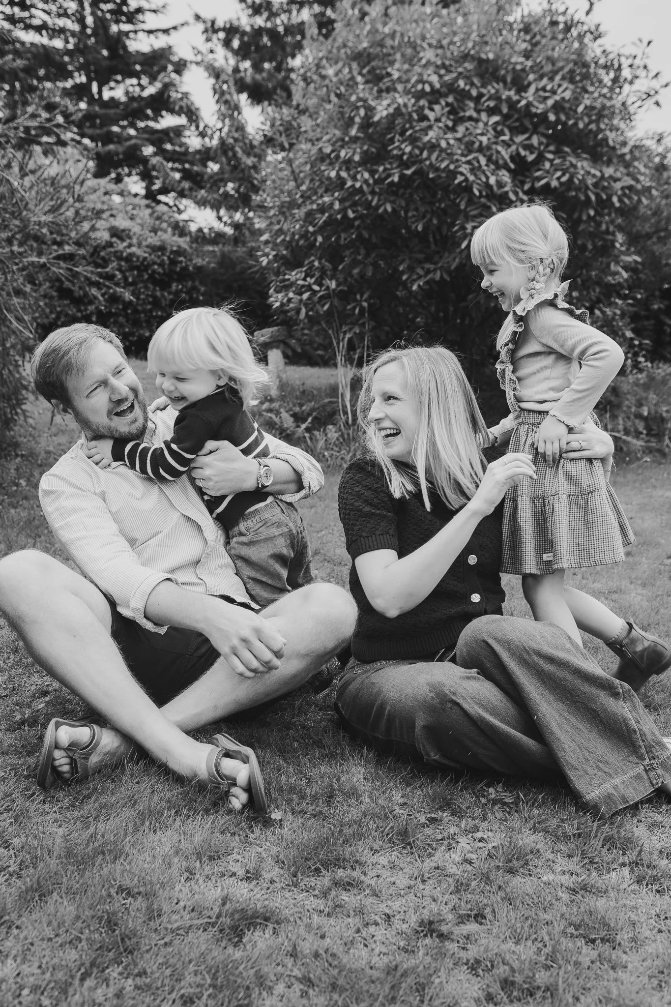 Black and white photo of a happy family outdoors, with a man, woman, and two young girls smiling and interacting on a grassy lawn with trees in the background.