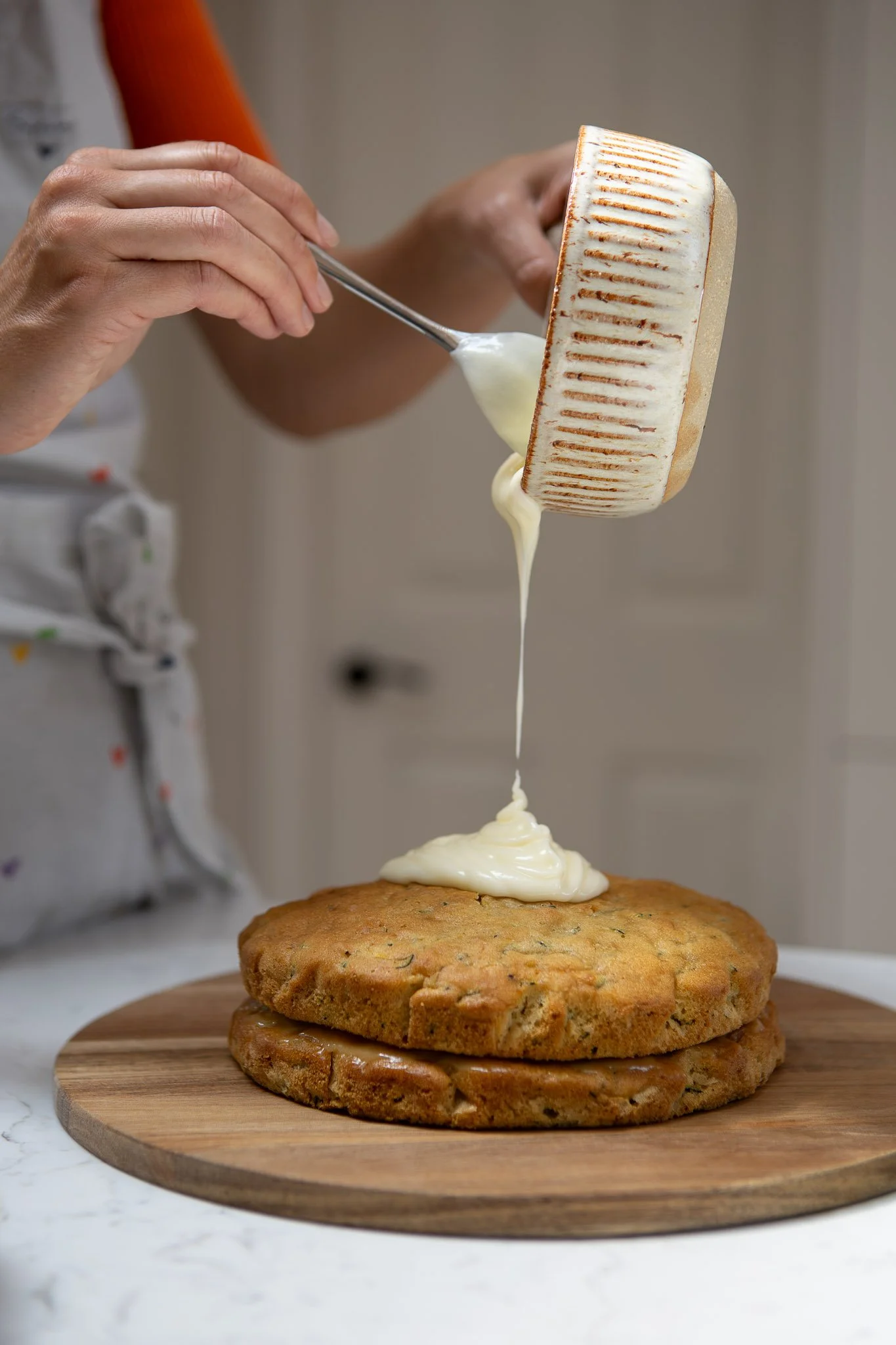 A person pouring white frosting from a spoon onto a layered cake on a wooden cutting board.