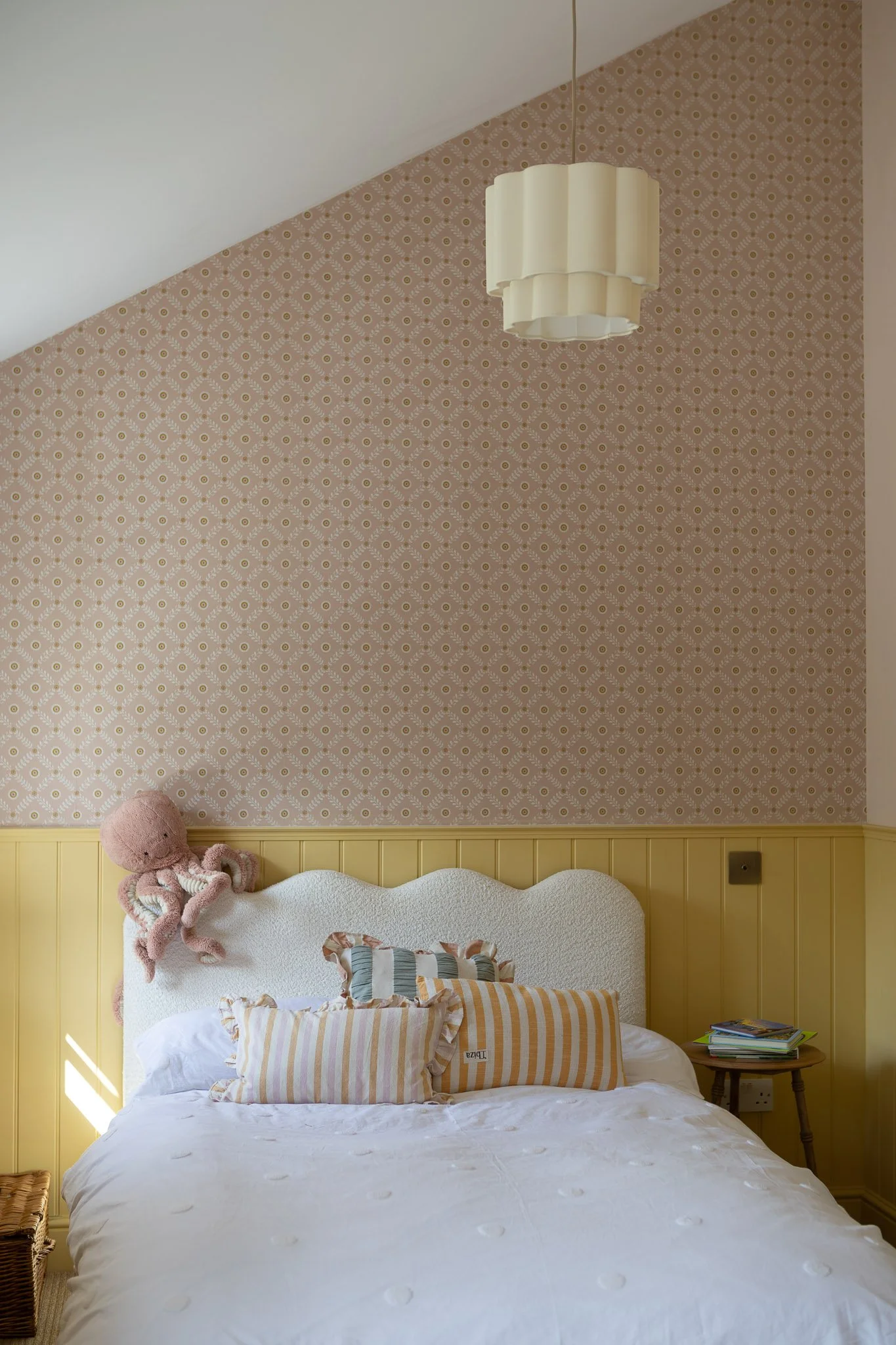 A cozy bedroom featuring a white bed with striped pillows, a yellow beadboard wall, a patterned pink wallpaper on the upper wall, a pink teddy bear, and a small wooden side table with books.