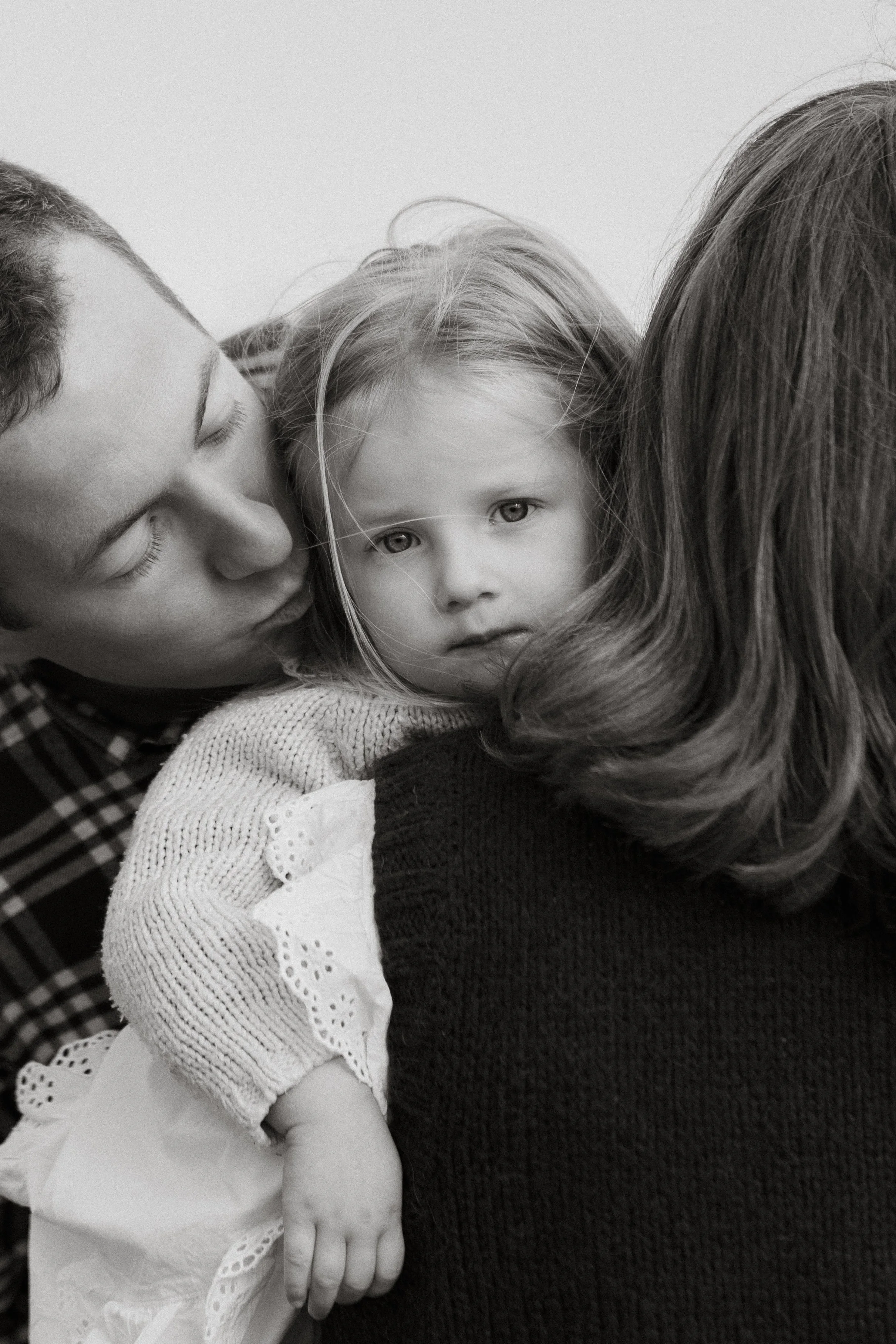 A woman with dark hair, a young girl with blonde hair and eyes, and a man with short hair, hugging each other closely.