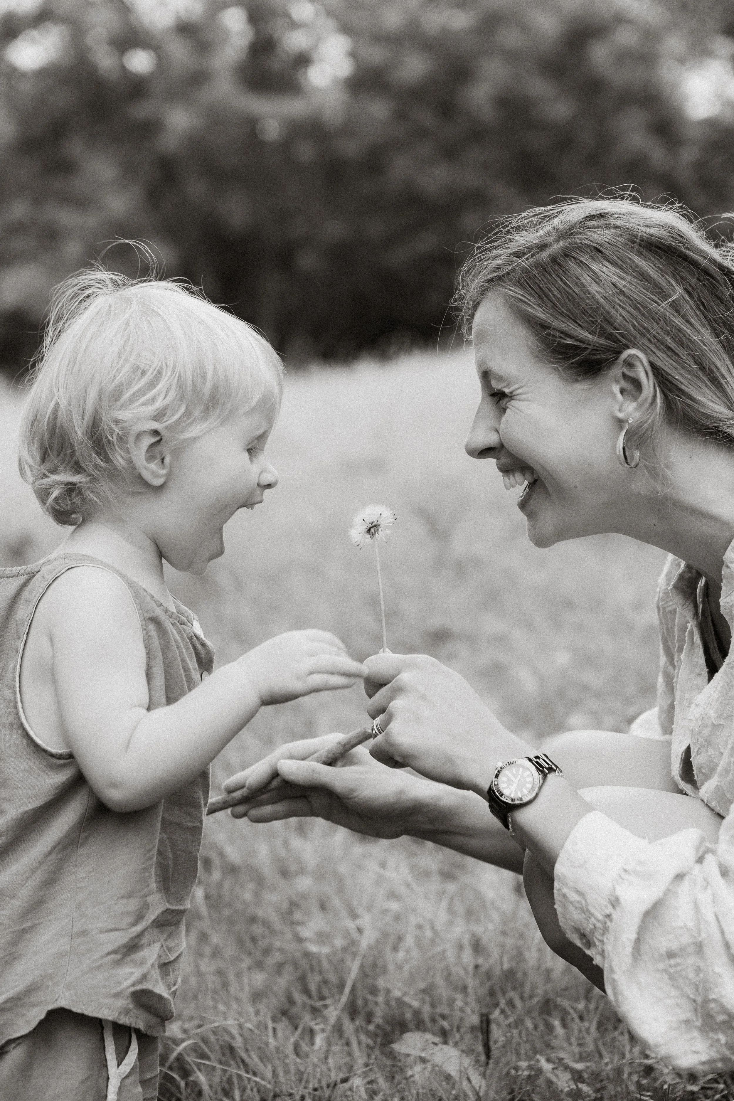 A woman and a young boy smiling and facing each other outdoors, holding a small flower together between them.
