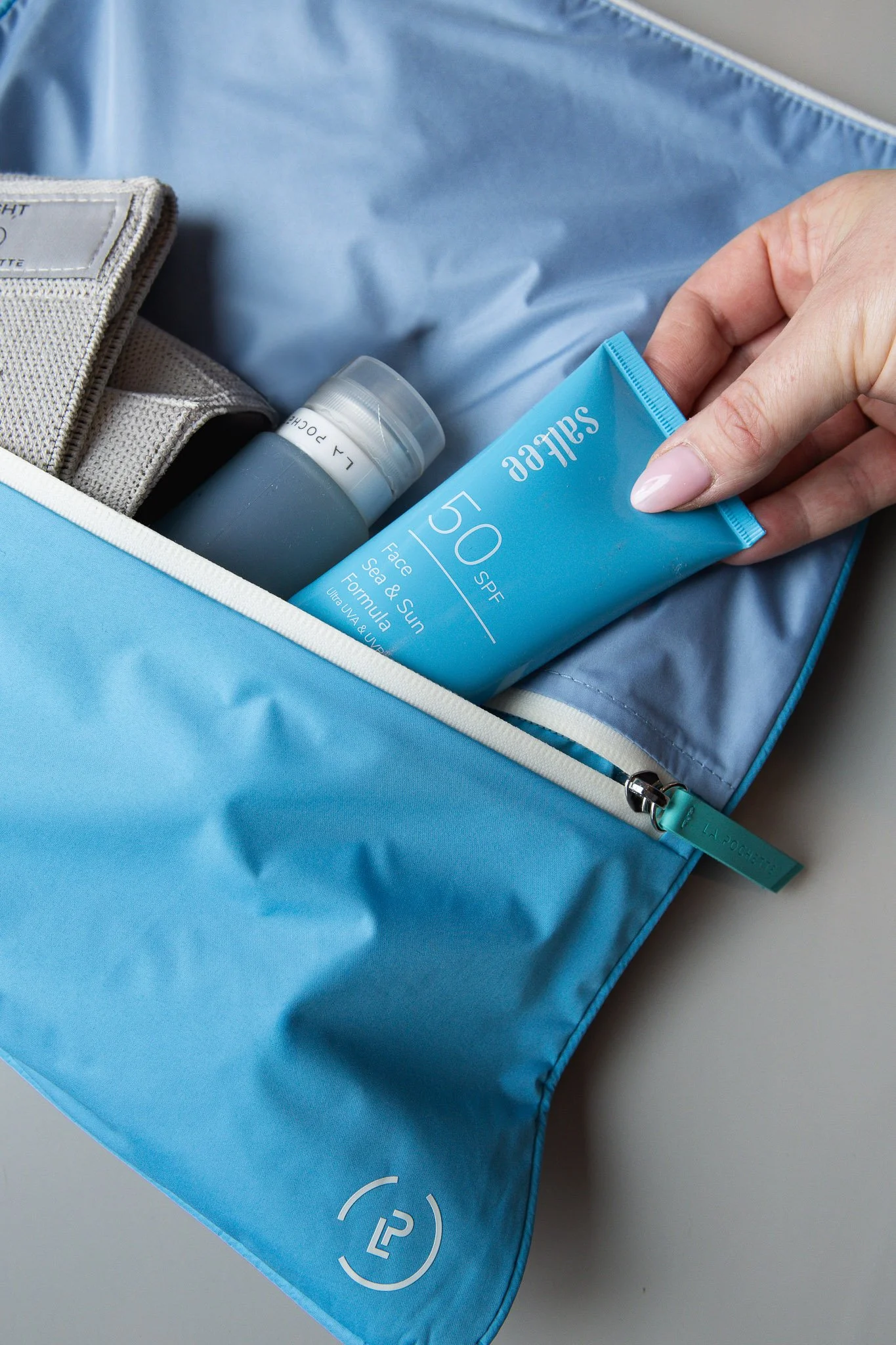 Open blue bag containing a gray water bottle, beige clothing, and a blue tube labeled SPF 50 sunscreen. The bag has a zipper with a blue pull tab.