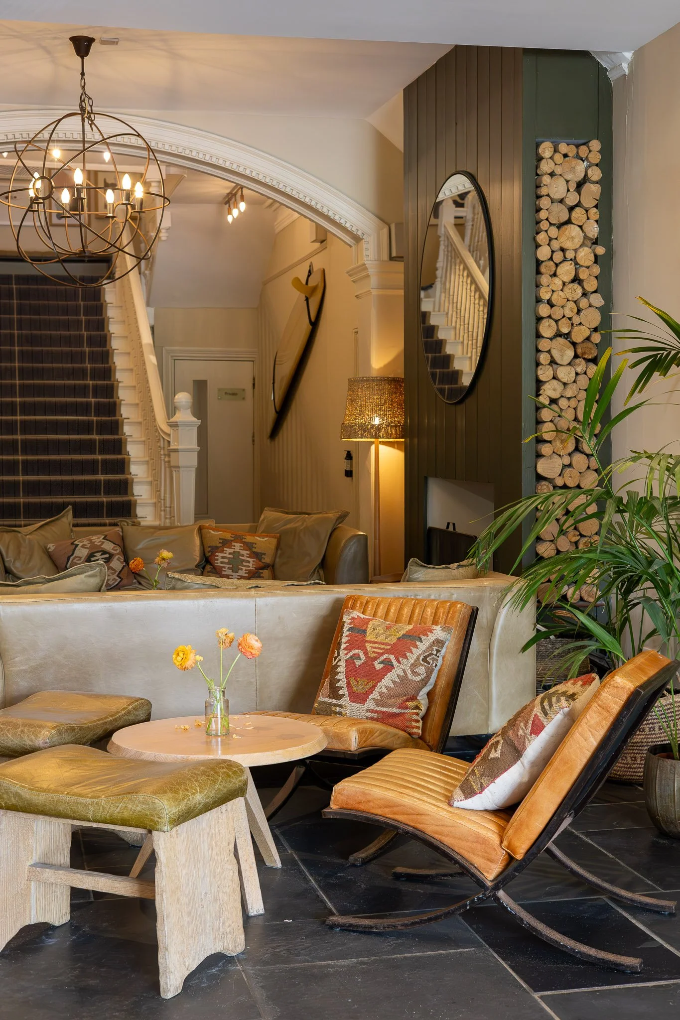 Interior living space with a staircase, hanging globe chandelier, sofa with pillows, round wooden table with a small flower arrangement, armchairs, standing lamp, and a wood-paneled wall with a mirror and a stack of firewood.