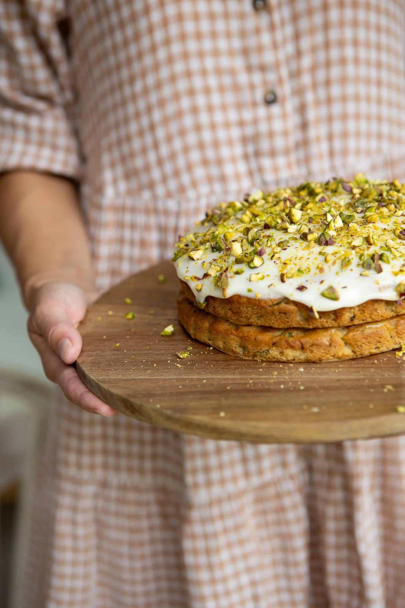 A person holding a wooden serving tray with a slice of cake topped with white frosting and chopped pistachios, wearing a checkered shirt.