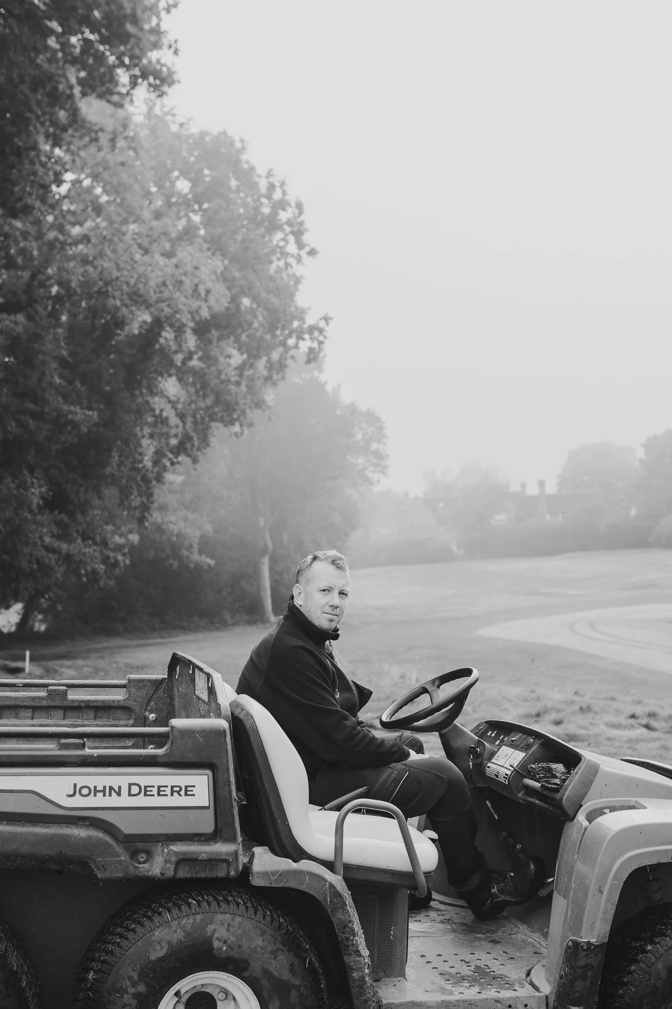 A man is sitting in a John Deere golf cart or utility vehicle in a park, with trees and a grassy area in the background.