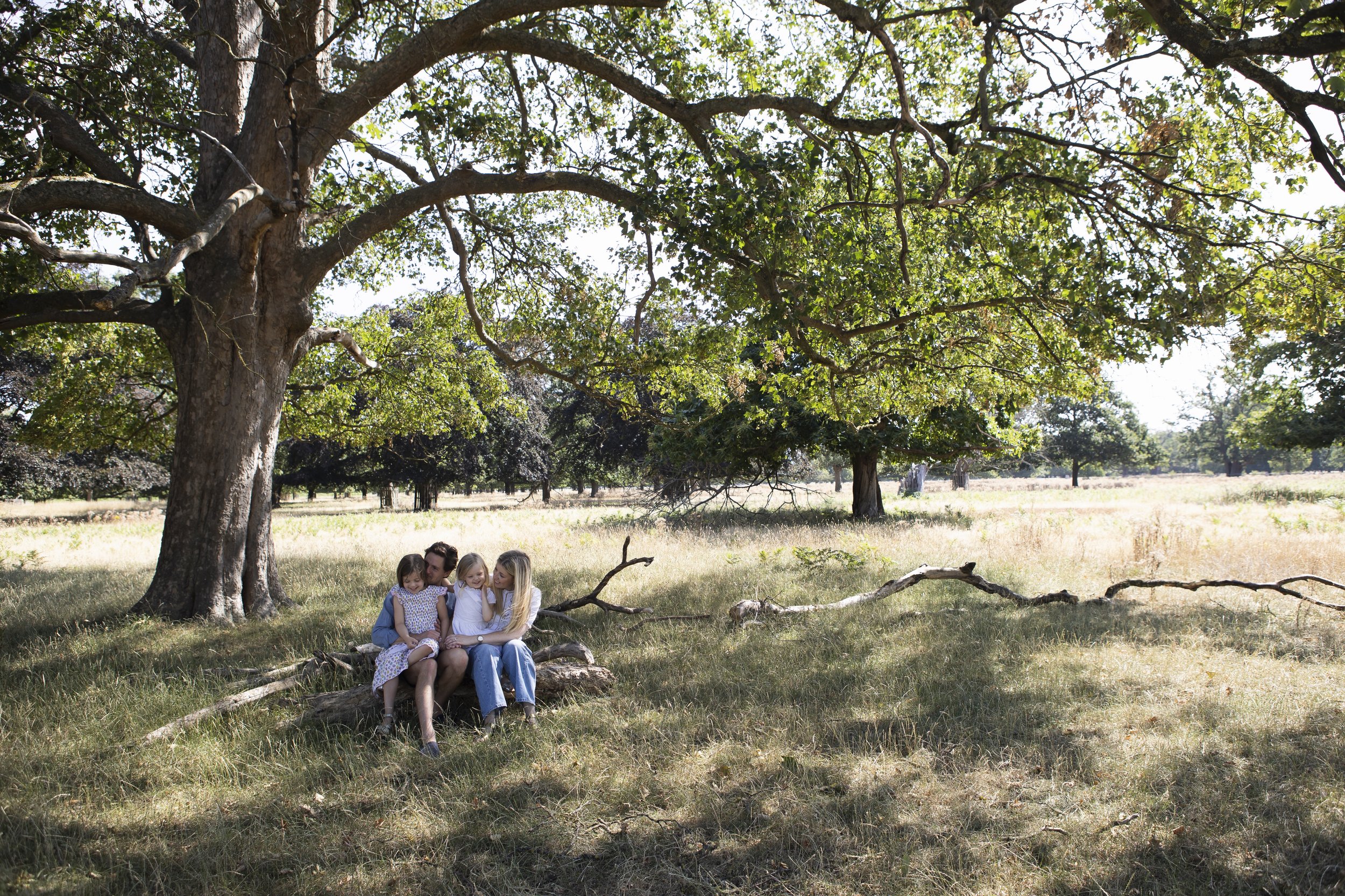 Two adult women and three young girls sitting together on the grass under a large tree in a park or countryside setting, with more trees and open field in the background.