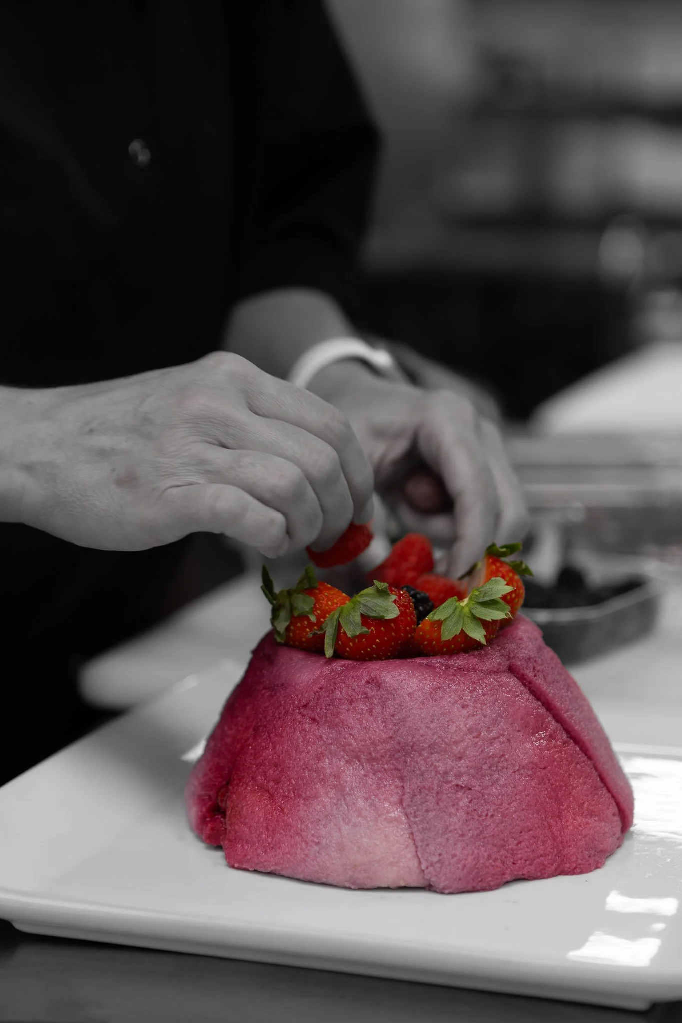 A pink mochi dessert topped with strawberries and other berries on a white plate, with a chef's hands in the background preparing the dessert.