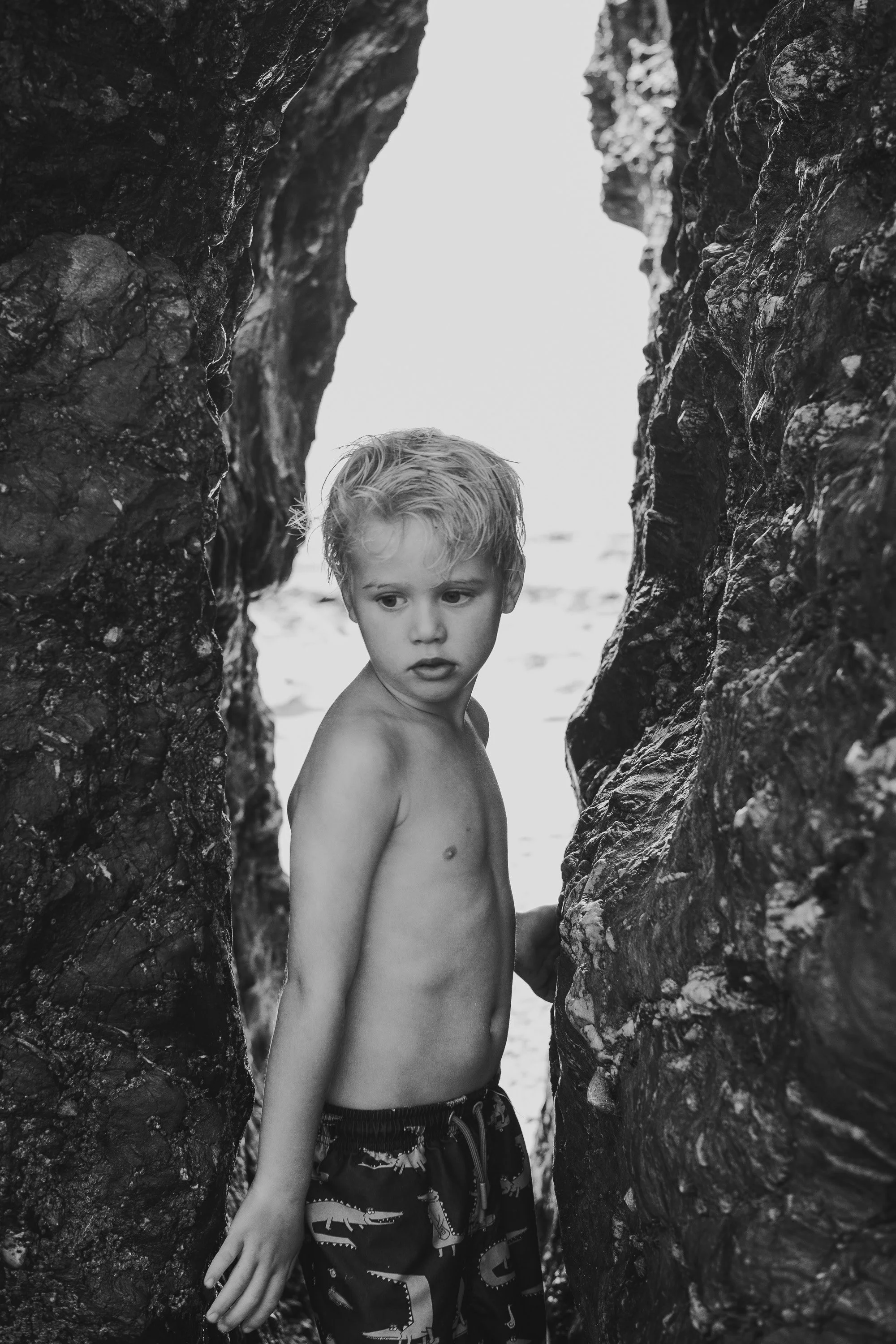 A young boy standing shirtless between two large, textured rocks with a bright sky in the background.