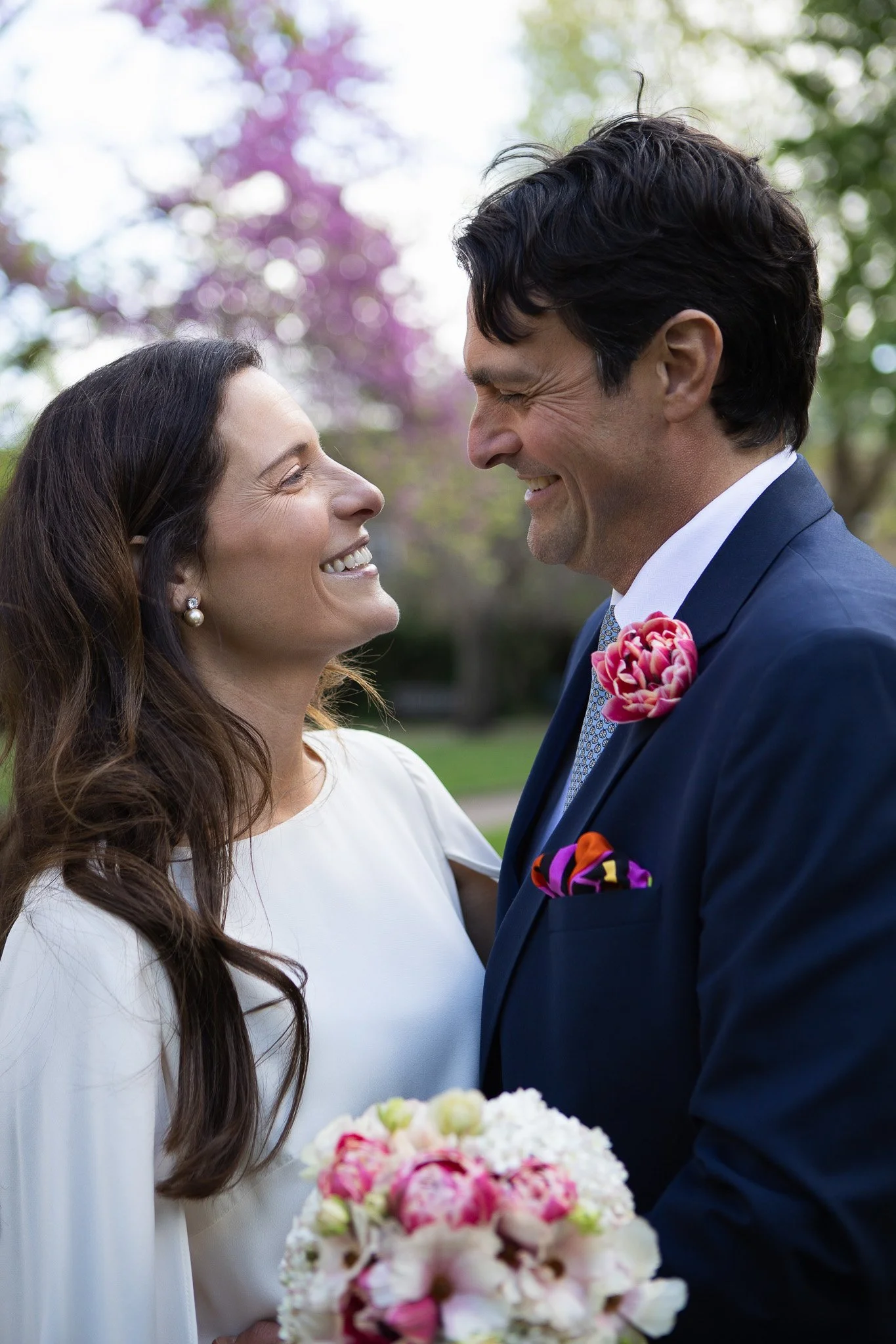 A smiling woman and man face each other closely, dressed in wedding attire, outdoors with flowers and trees in the background.