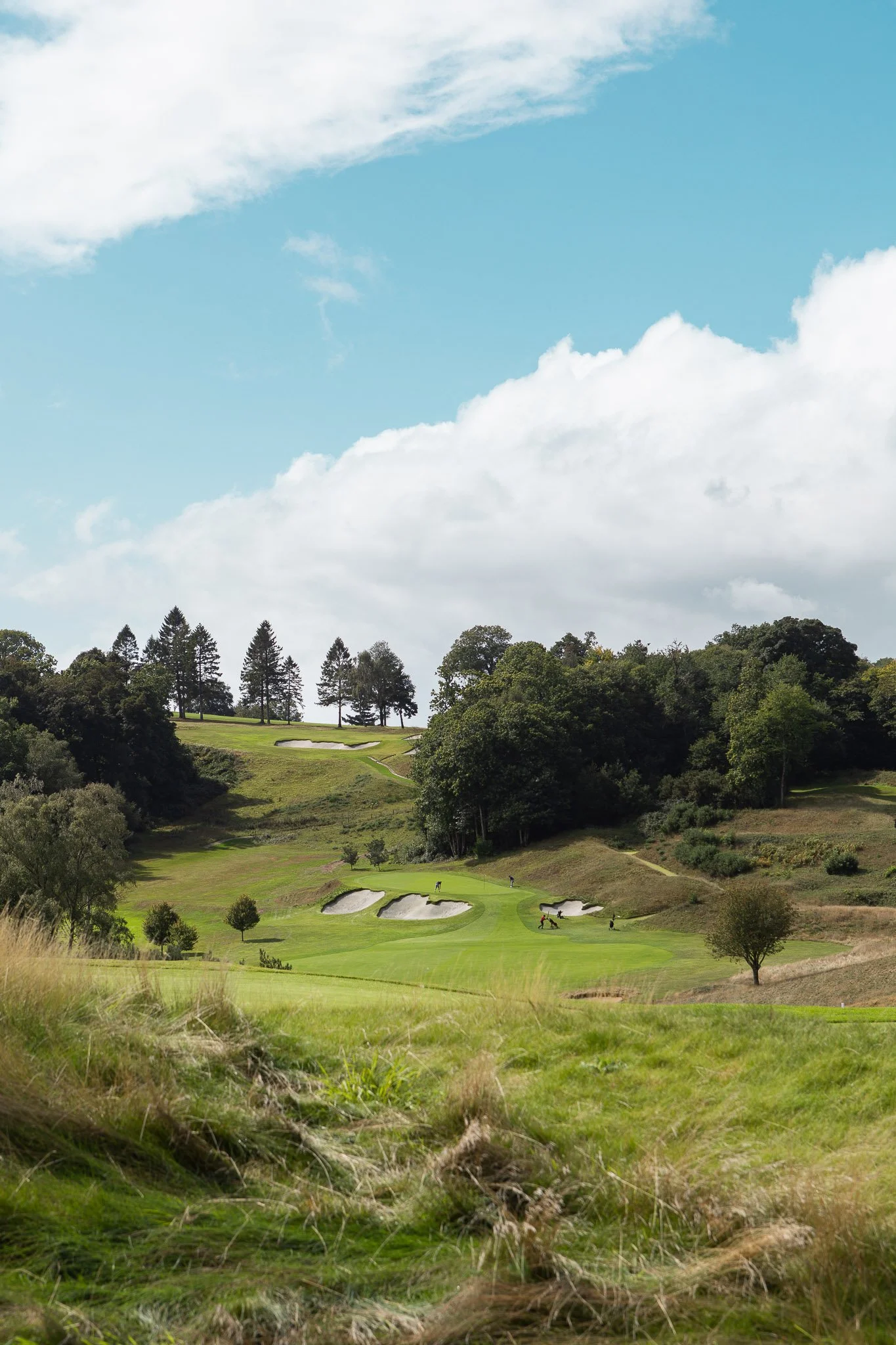 A scenic view of a golf course surrounded by lush green trees with a few players and sand traps, under a bright blue sky with scattered clouds.