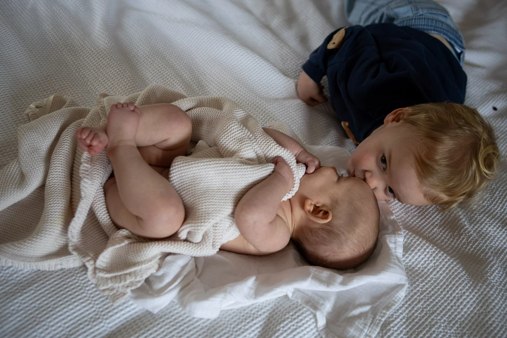 A person with a baby on a bed, with a young child leaning over and kissing the baby's forehead.