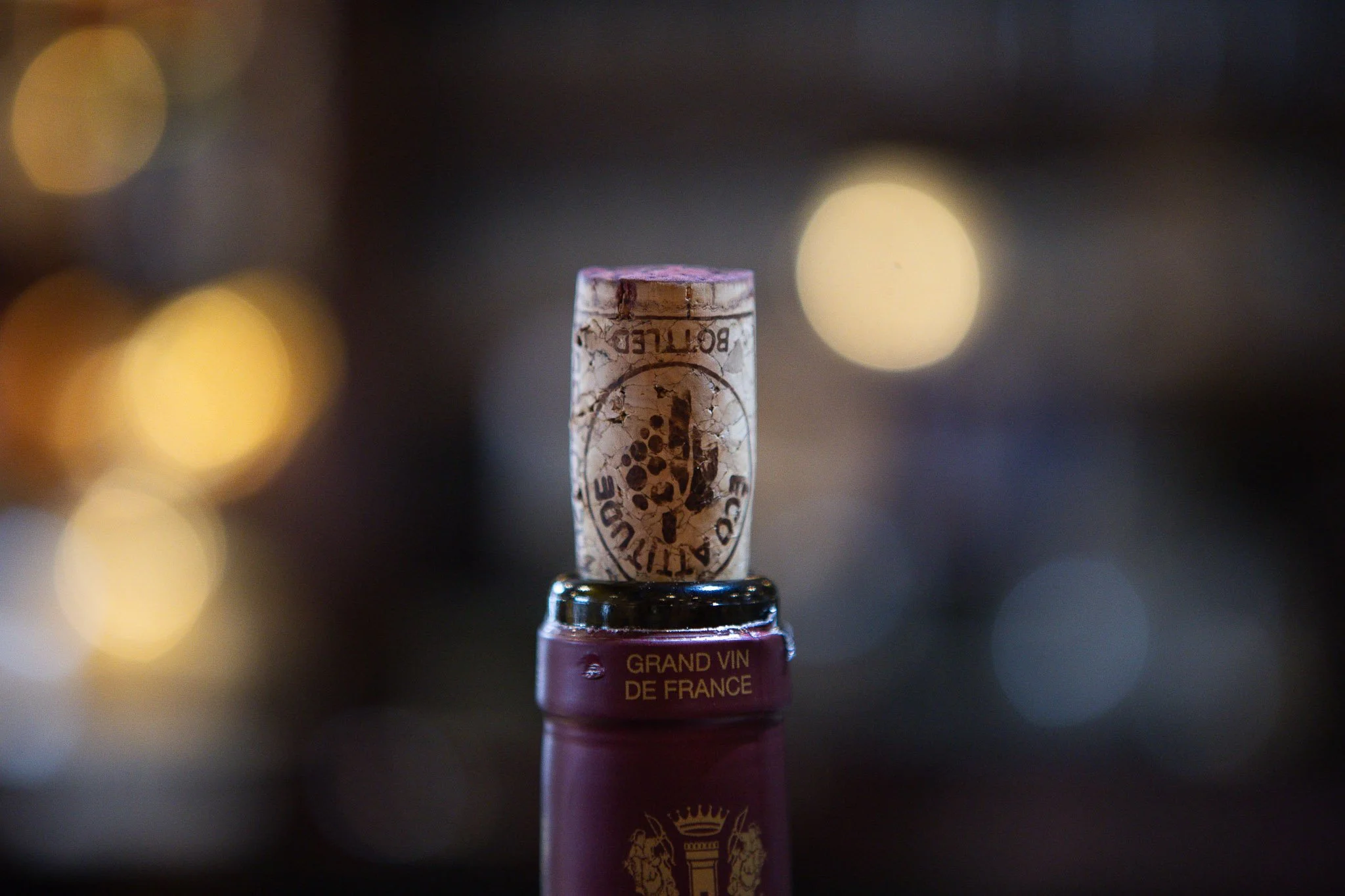 Close-up of a wine cork with text and logo, inserted into a wine bottle with a blurred background of warm lights.