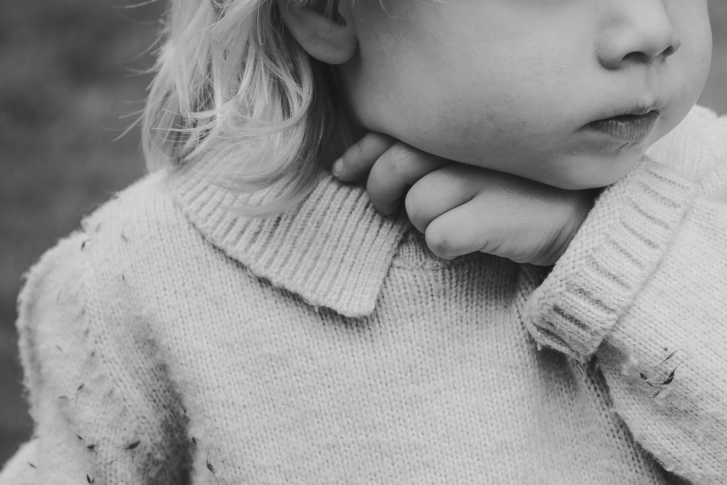 Close-up of a young child's face and hand, with blonde curly hair, wearing a knitted sweater, in black and white.