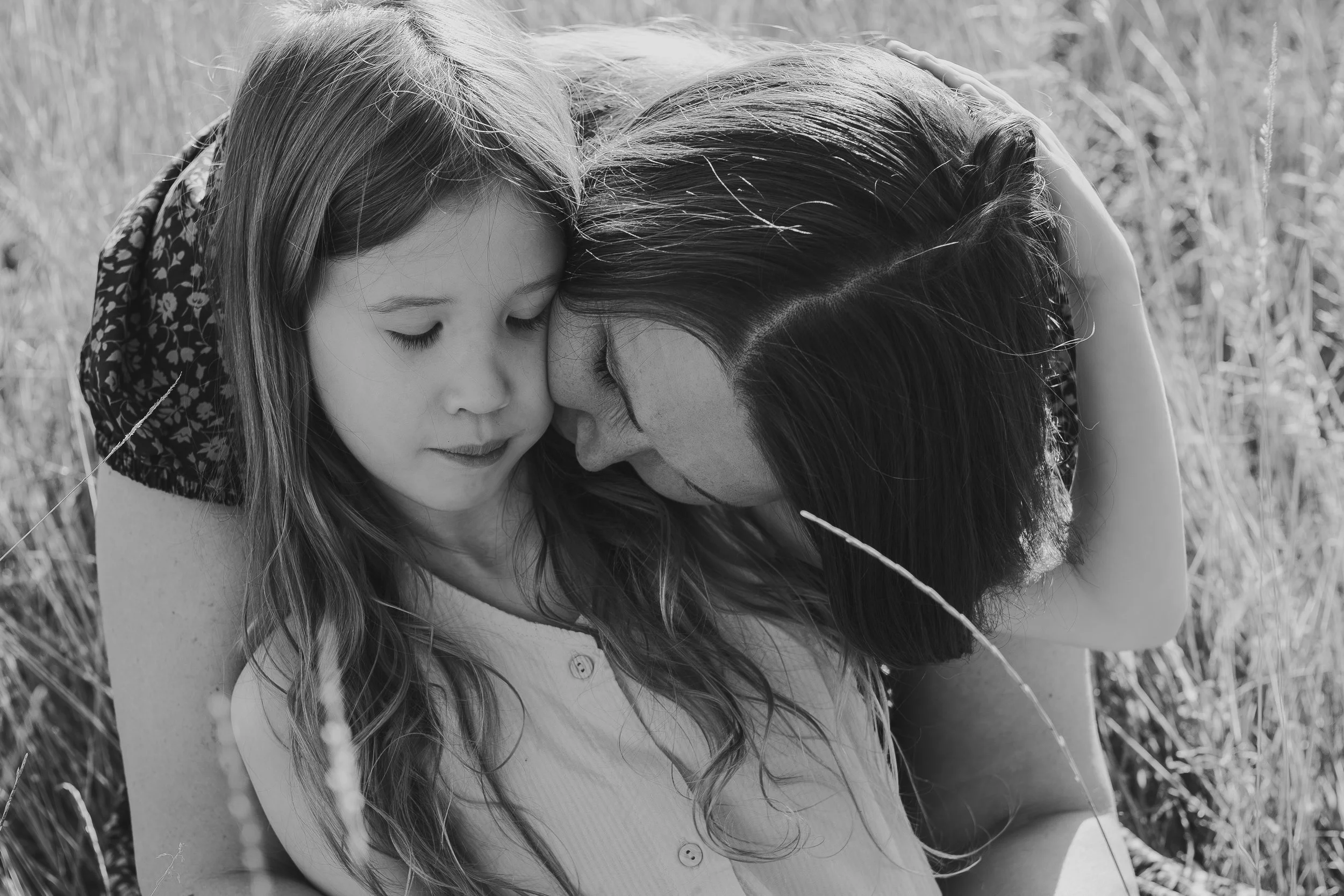 A woman and a young girl gently touching their foreheads together with closed eyes in a grassy outdoor setting.