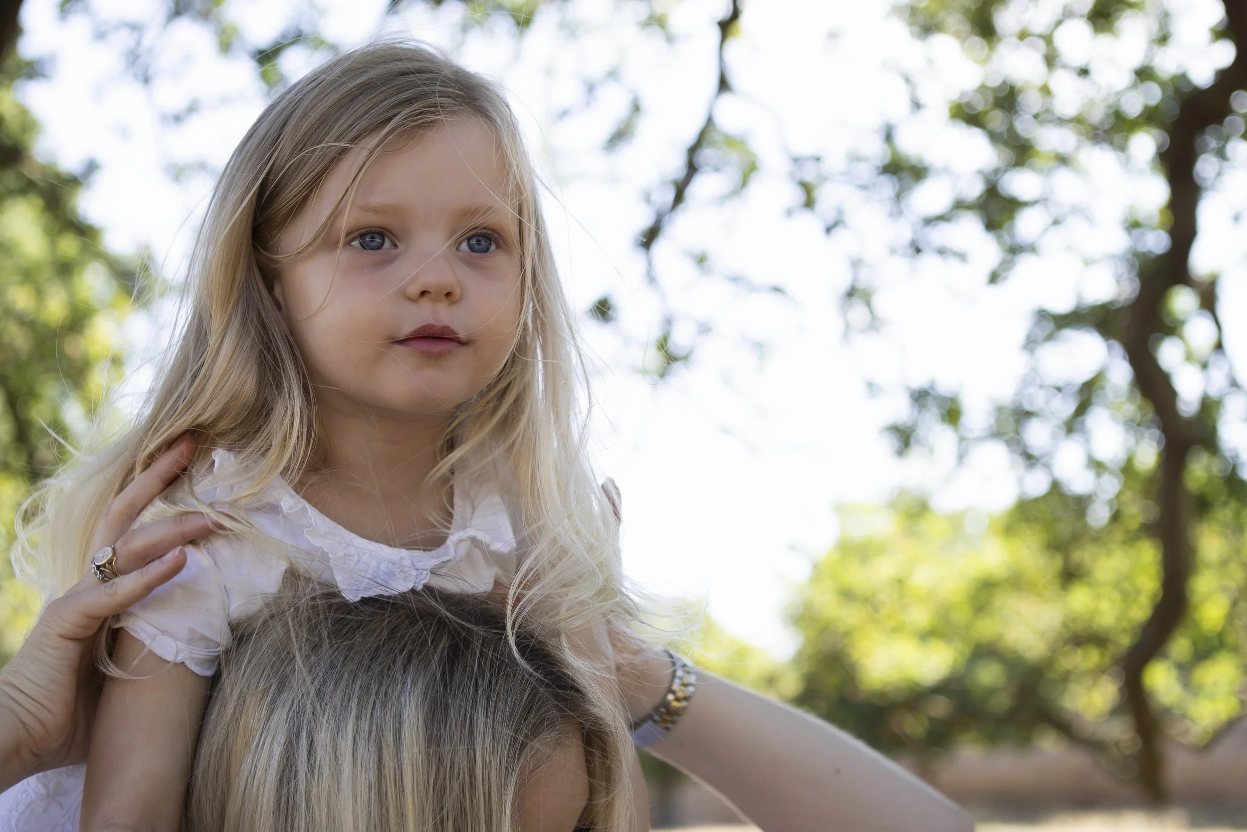 A young girl with long blonde hair and blue eyes, wearing a white shirt, being carried on someone's shoulders outdoors among trees with sunlight filtering through the leaves.