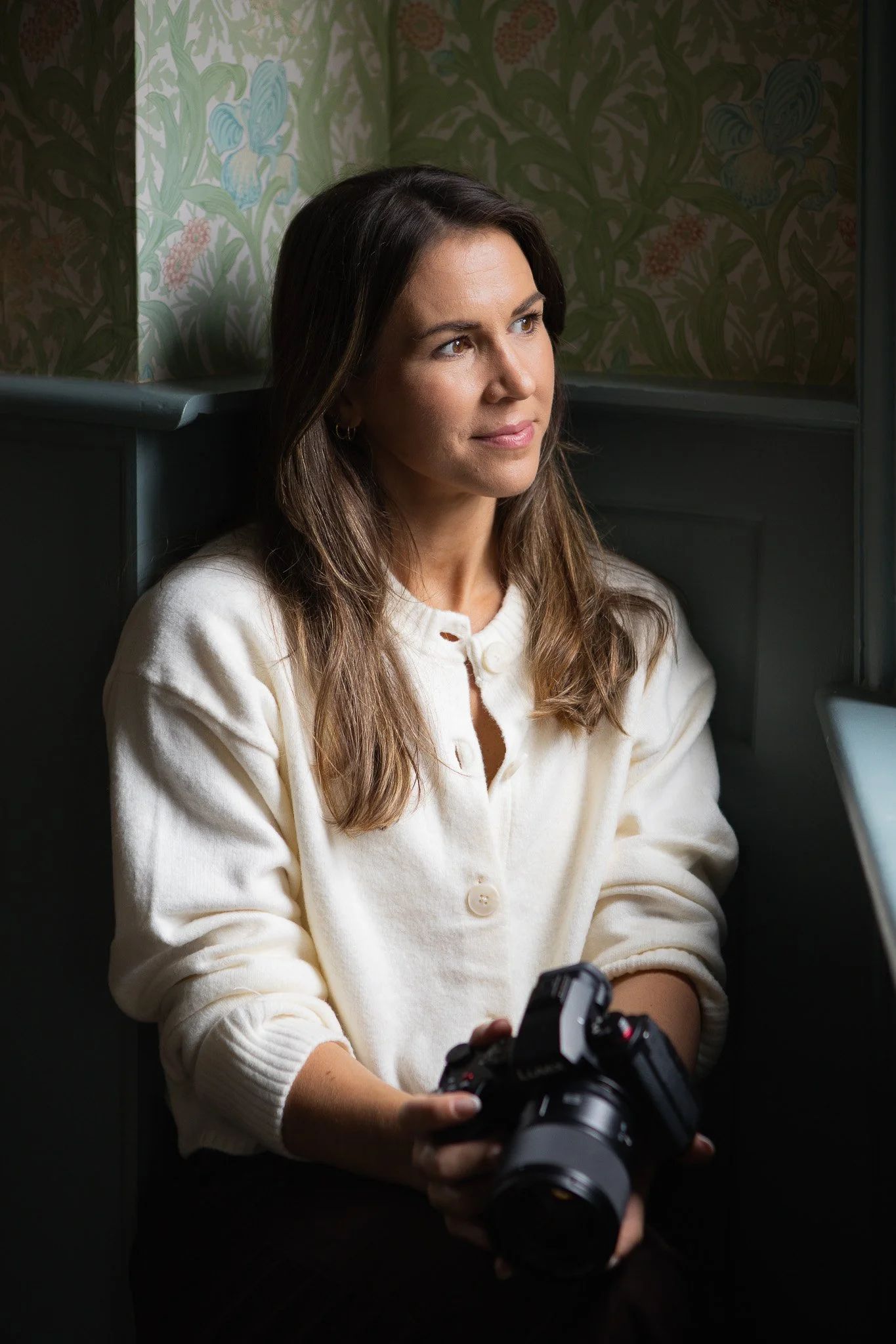 A woman with long brown hair and light skin, wearing a cream-colored sweater, holding a camera in her hands. She appears to be sitting indoors with a wallpapered background featuring blue and pink floral and butterfly patterns.