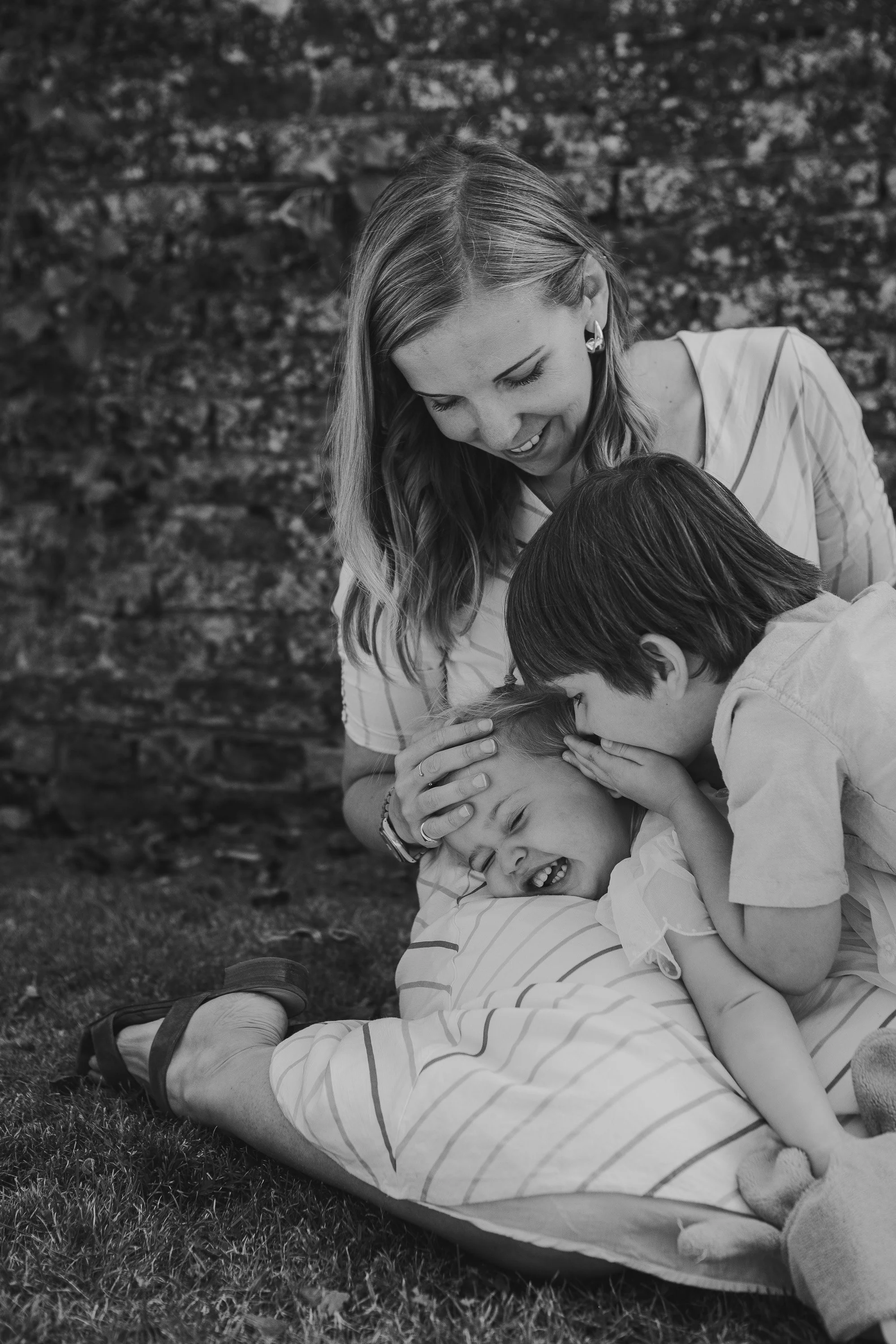 A woman and two children playing on the grass, with the children laughing and the woman smiling, in front of a stone wall.