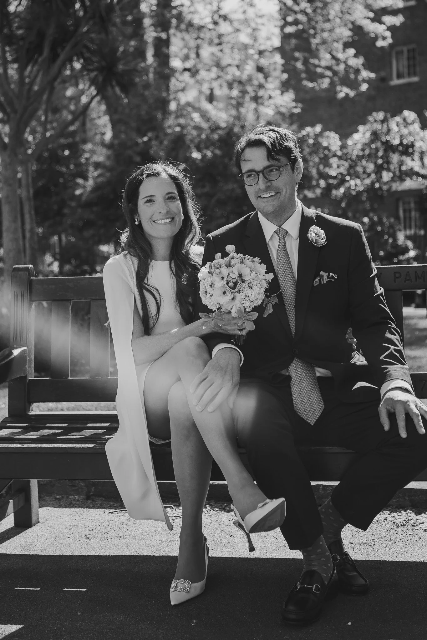 A black-and-white photo of a couple sitting on a bench outdoors, smiling and holding a bouquet of flowers. The woman is wearing a dress and high heels, while the man is dressed in a suit with glasses. They are surrounded by trees and a building in the background.