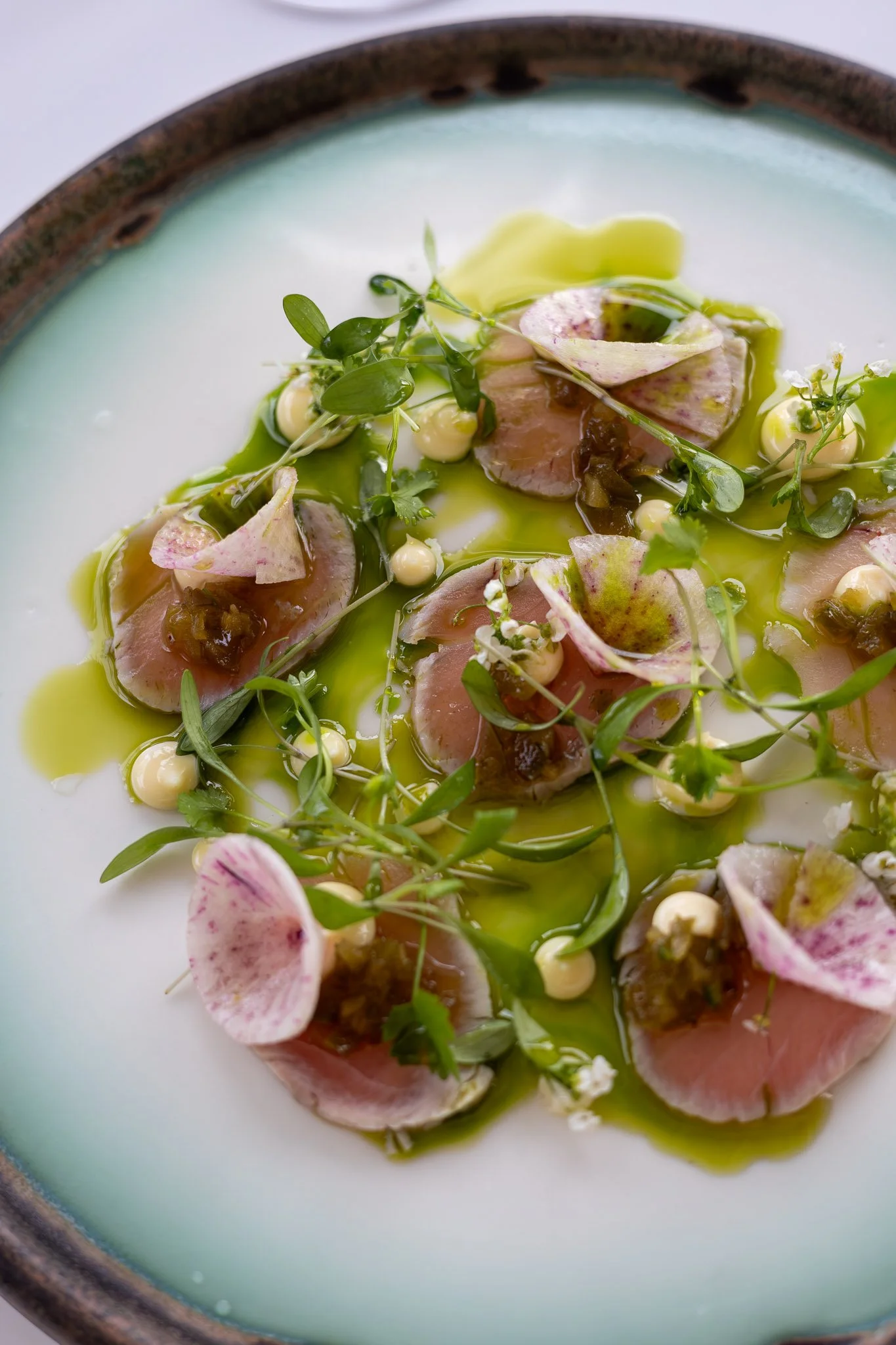 Close-up of a plated dish featuring sliced raw fish, microgreens, edible flowers, small dollops of sauce or mousse, and a green oil or sauce drizzle on a ceramic plate with a dark rim.