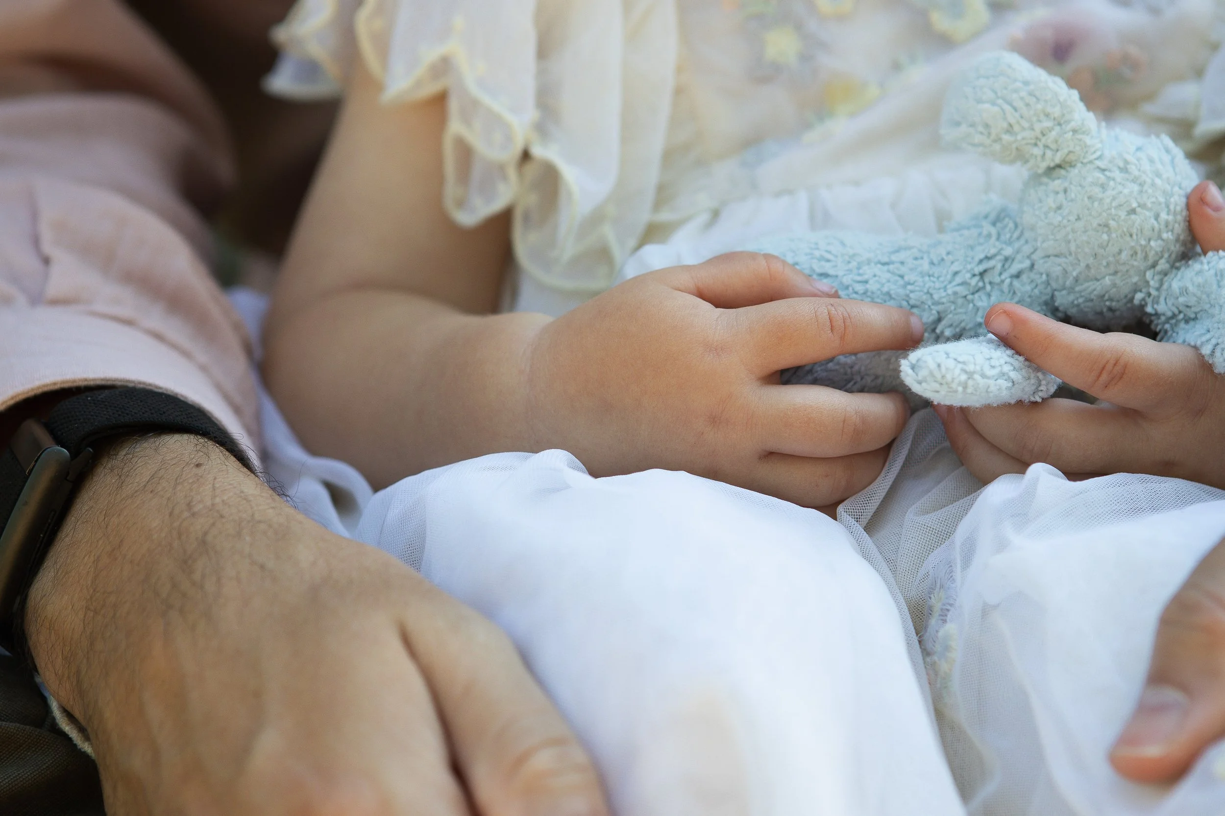 A child holding a stuffed animal, possibly a plush bear, while laying on an adult's arm. The child's arm is visible, and they are wearing a light-colored, ruffled garment. The adult's arm, which is around the child, is partially visible on the left s
