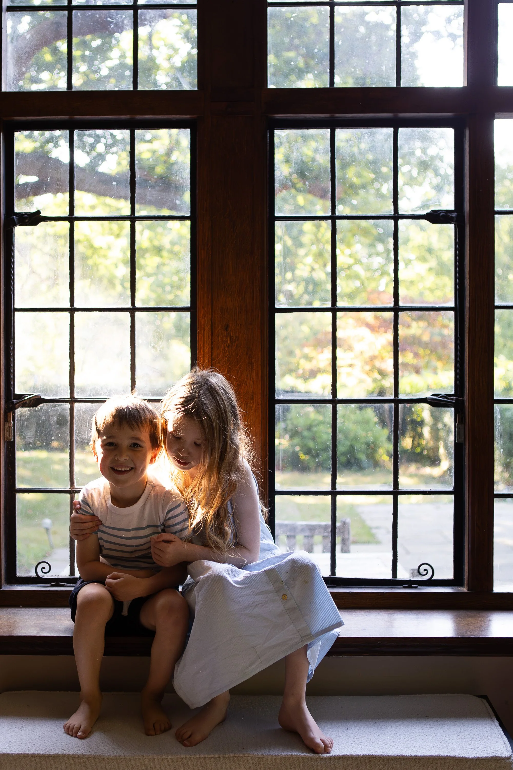 Two children, a boy and a girl, sitting on a window sill inside a house. The boy is smiling at the camera, and the girl is looking at him with a smile. They are near a large wooden window with multiple panes, with sunlight and trees visible outside.