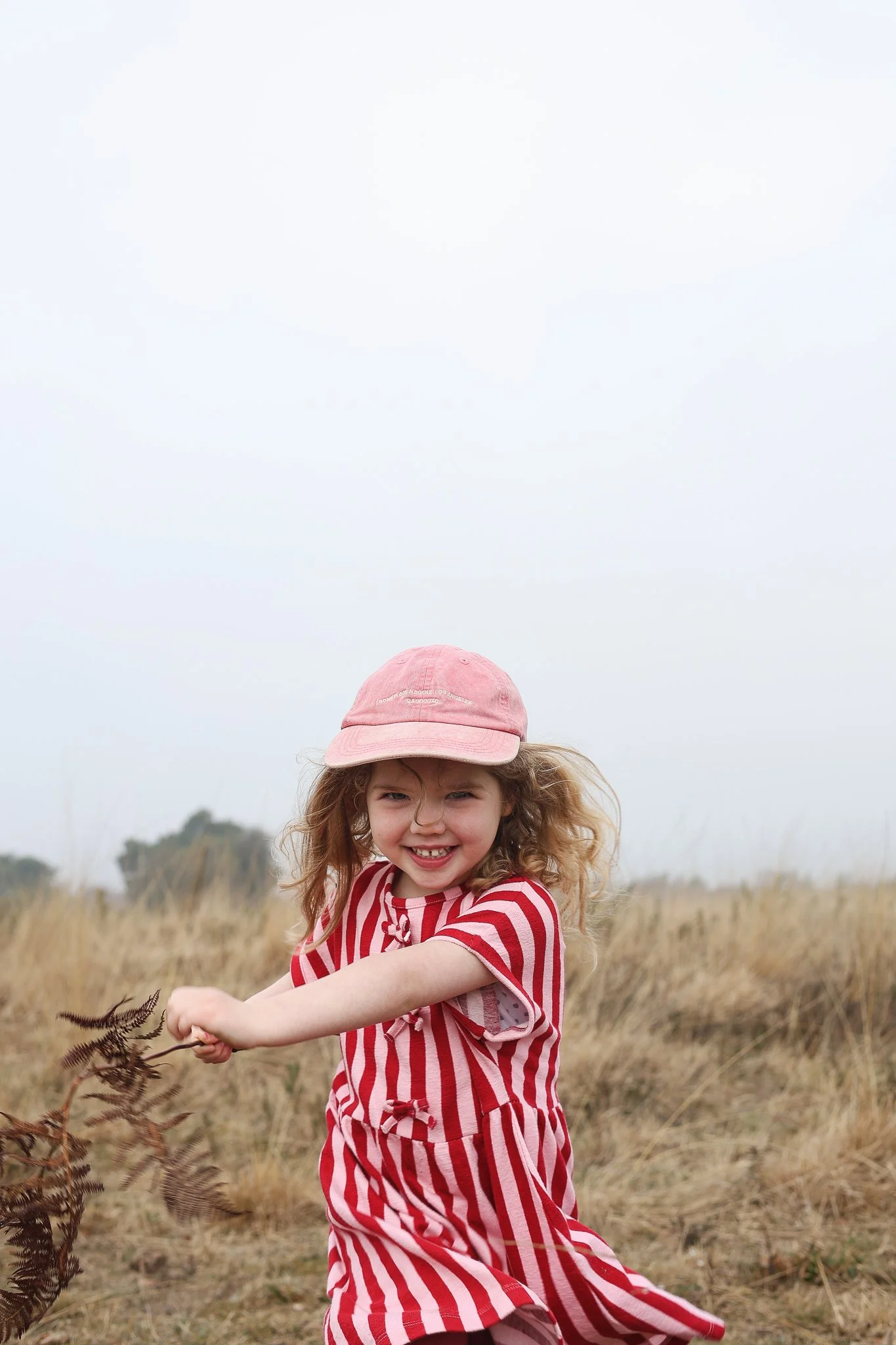 A young girl with curly hair wearing a pink cap and a red and white striped dress, smiling and sitting in a field of tall grass during daytime.