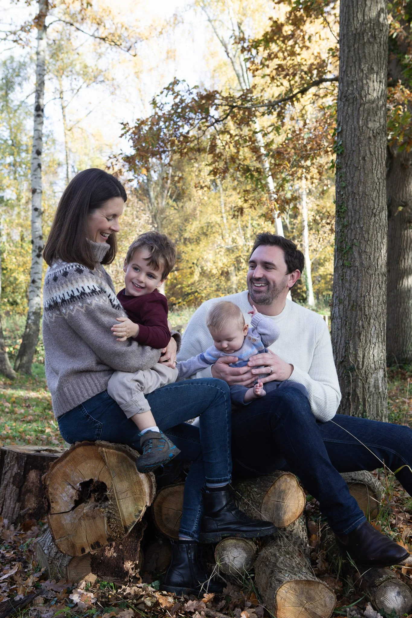 A family of five enjoying time outdoors in a wooded area with fall foliage, sitting on a large chopped tree log, smiling and interacting.