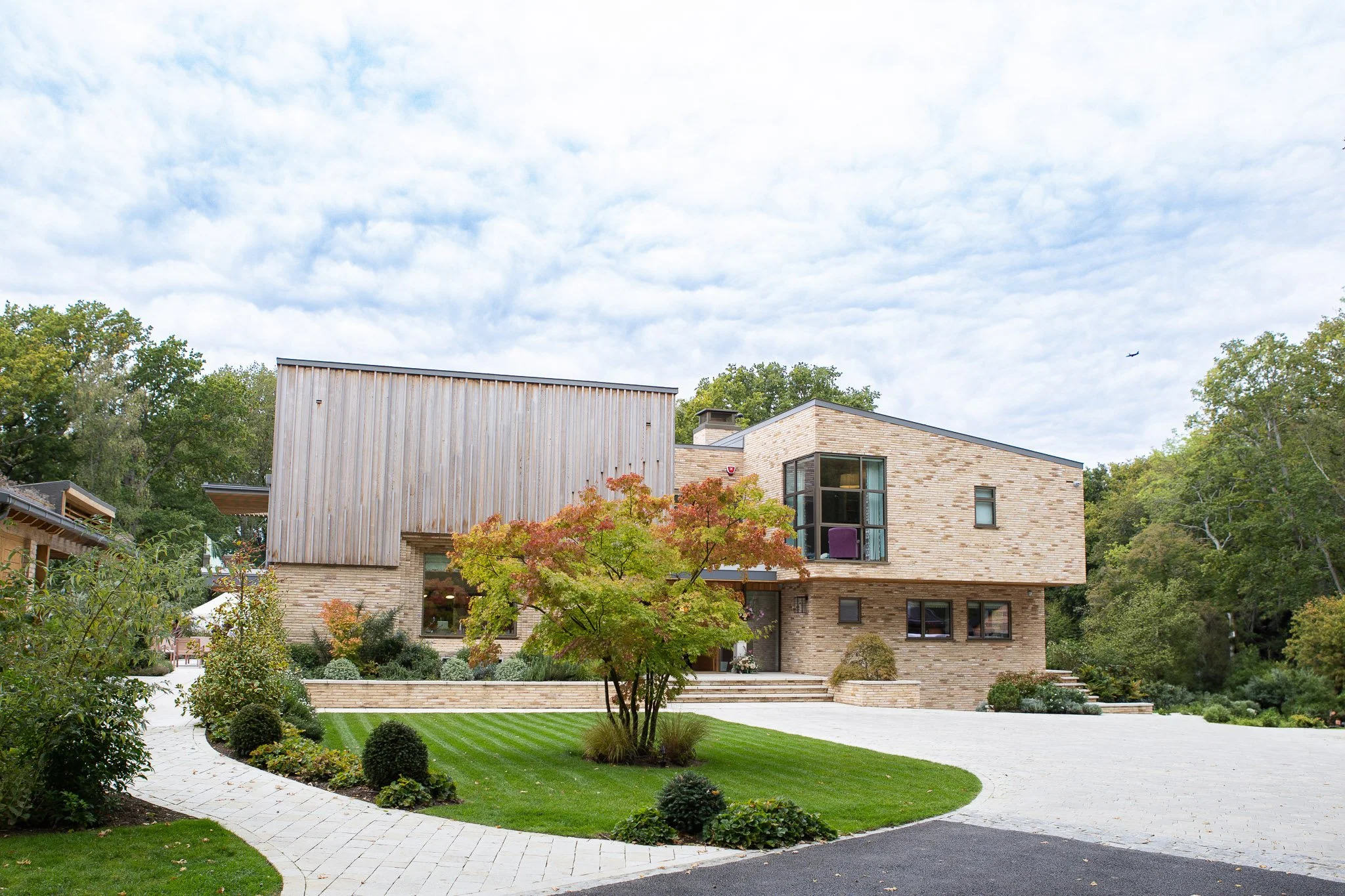 Modern house with landscaped front yard, curved driveway, green lawn, trees, and cloudy sky.