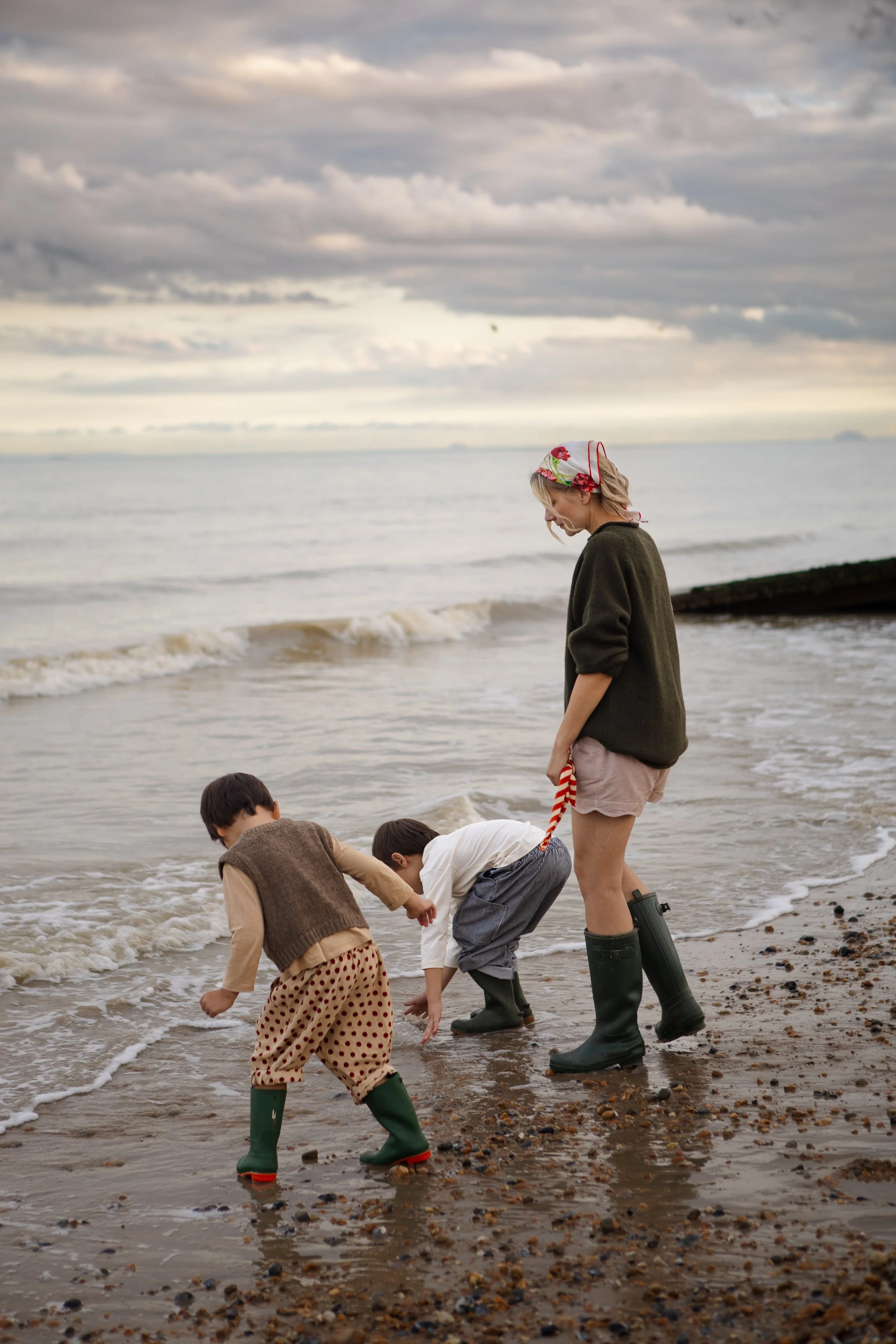 A woman and three children wearing rubber boots, playing and exploring at the beach during cloudy weather, with waves and rocks in the background.