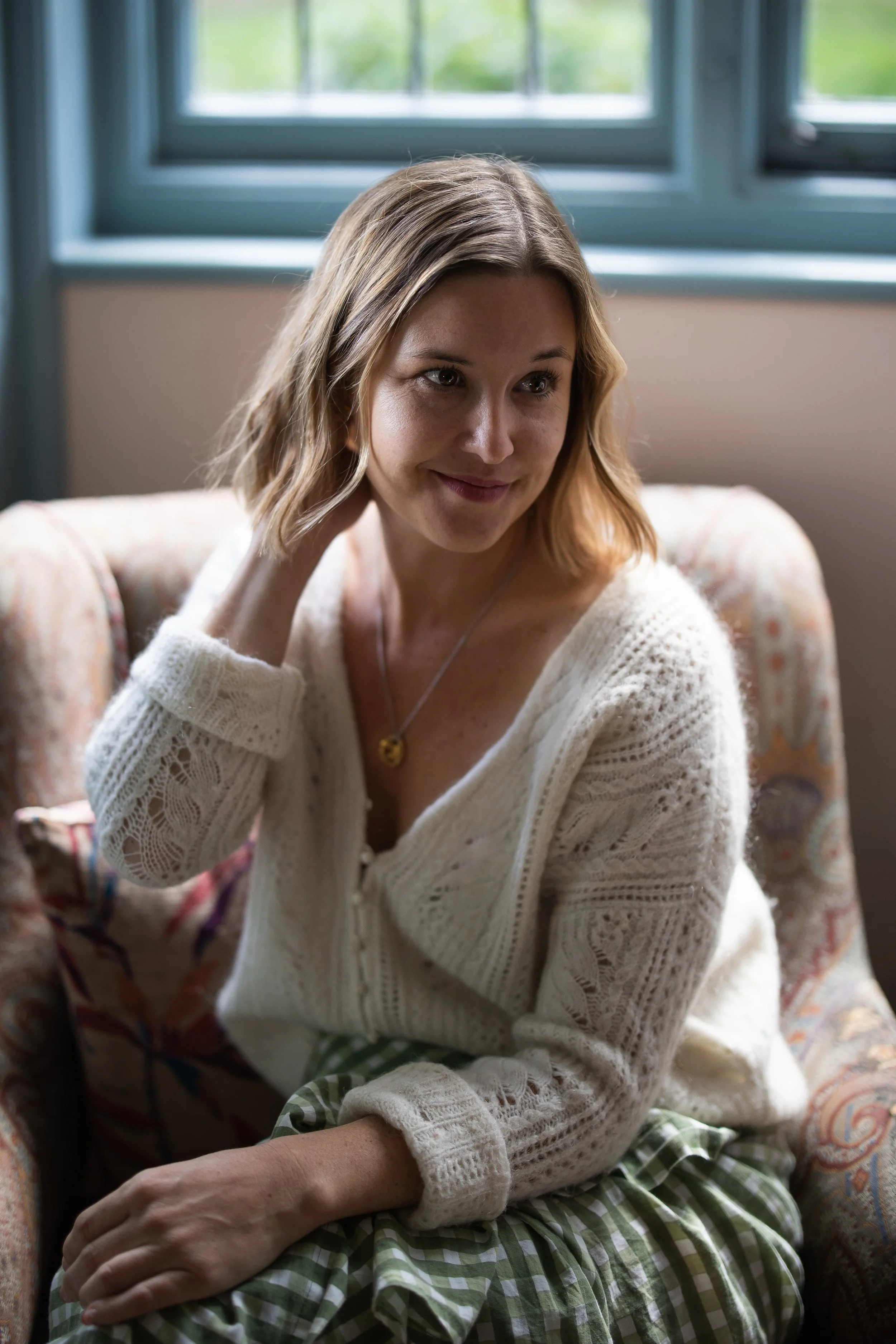 A woman with shoulder-length blonde hair smiling and looking slightly to the side, wearing a white knitted sweater and sitting on a patterned armchair, with a large window with blue trim in the background.