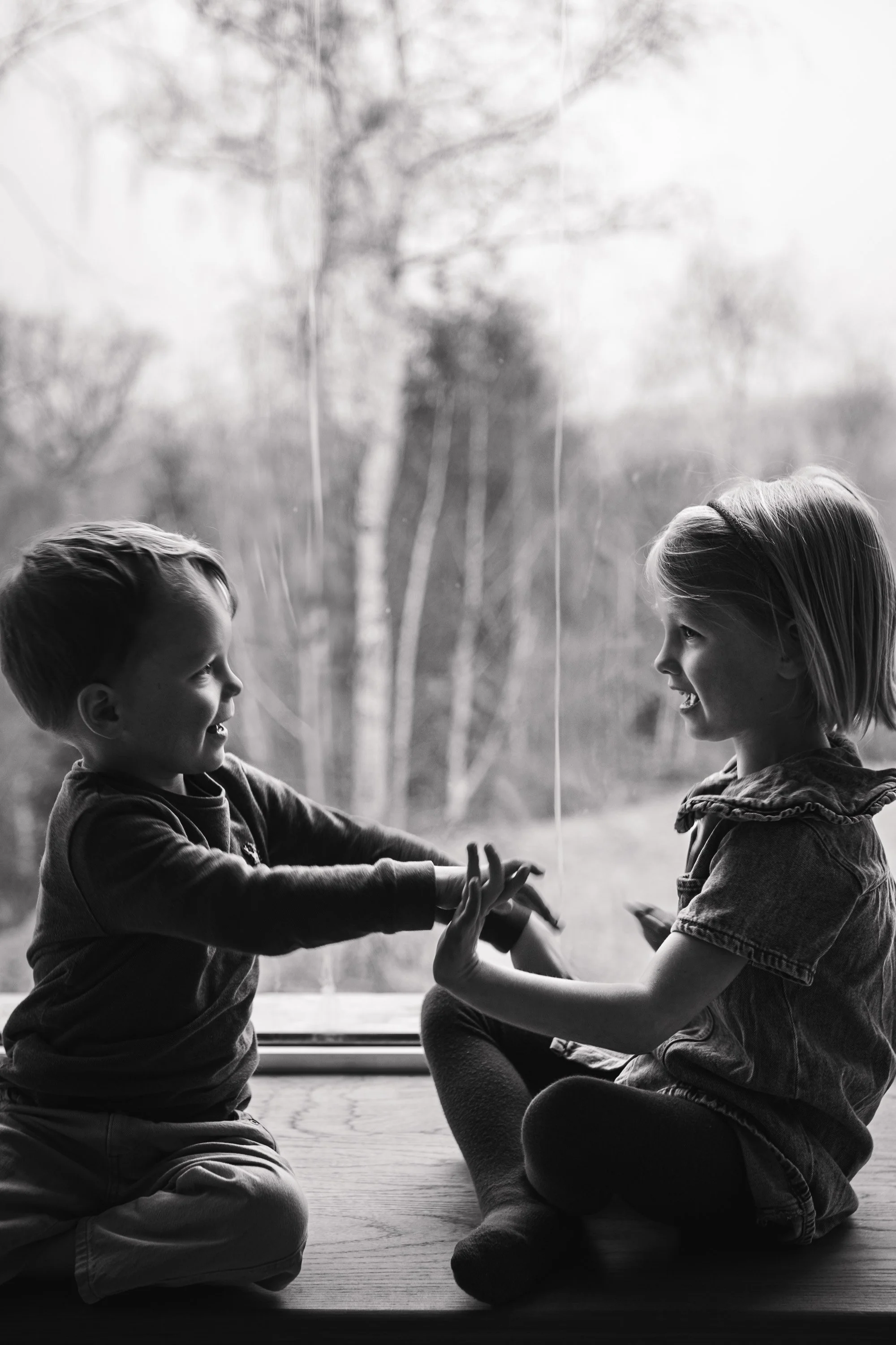 Two children, a boy and a girl, are sitting on the floor facing each other and playing together, holding hands, near a window with blurred outdoor trees in the background.