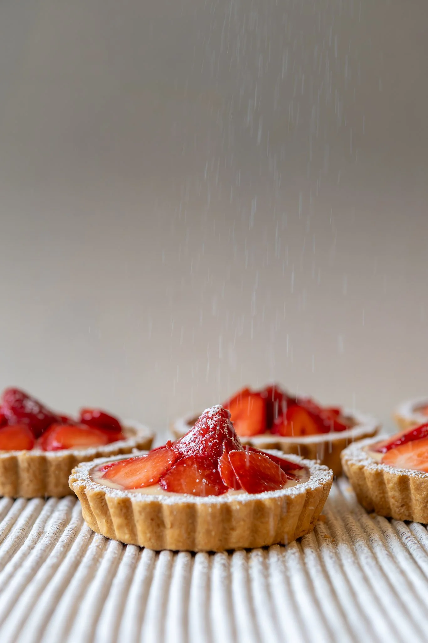 Close-up of strawberry tarts topped with sliced strawberries and a dusting of powdered sugar, on a textured surface.