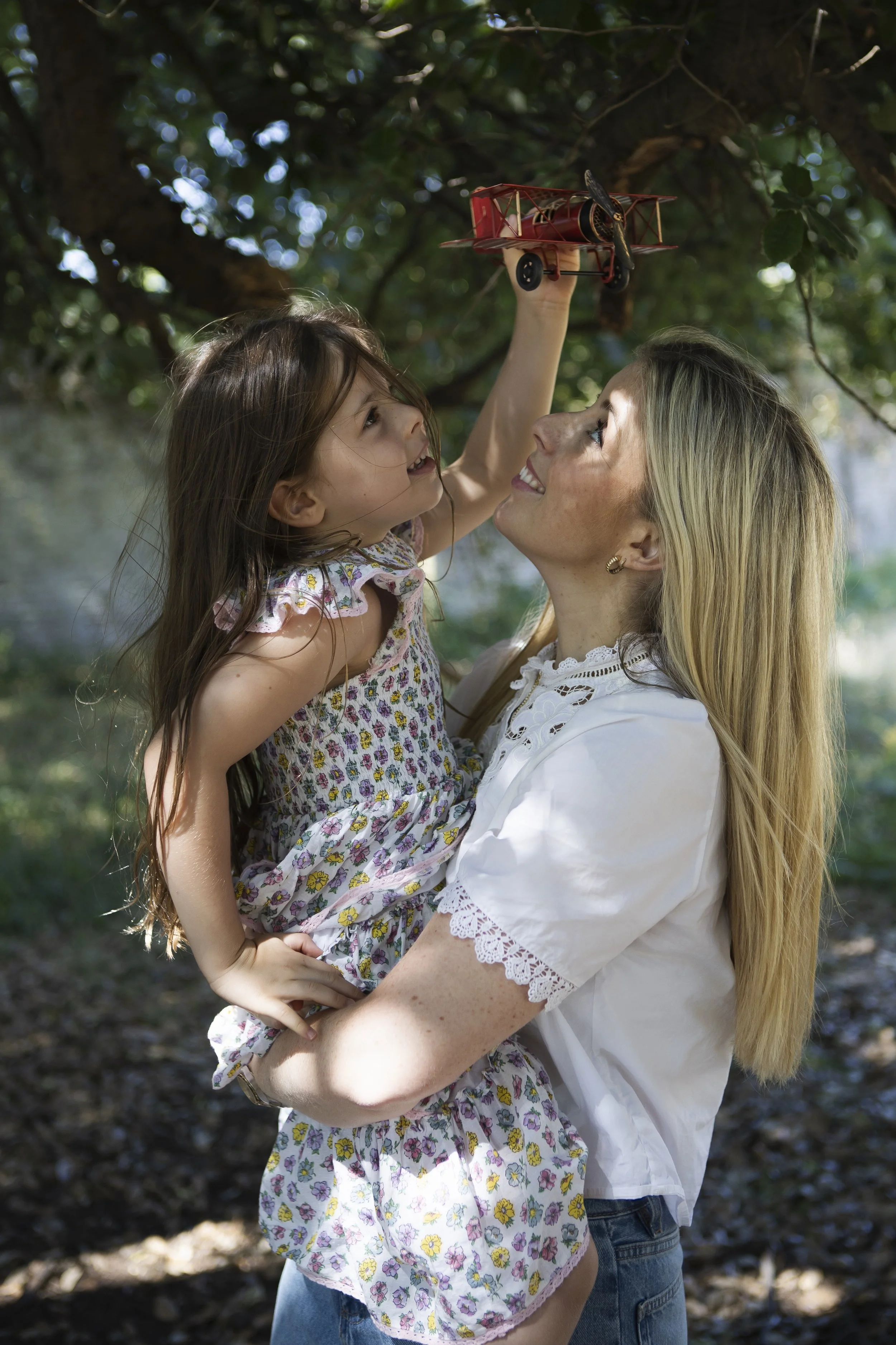 A woman holding a young girl in her arms outside among trees, playing with a red toy biplane, both smiling and looking at each other.