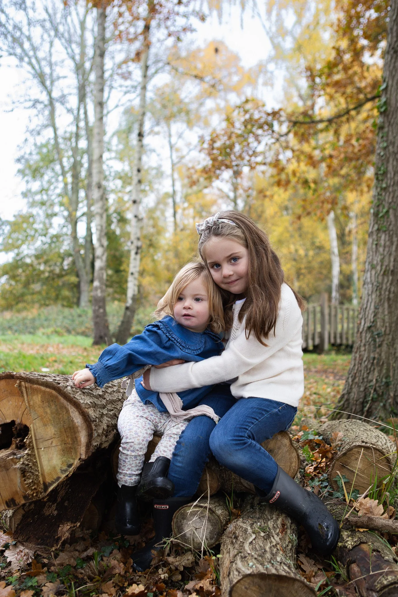 Two young girls sitting on a fallen tree trunk outdoors during fall, surrounded by trees with autumn-colored leaves.