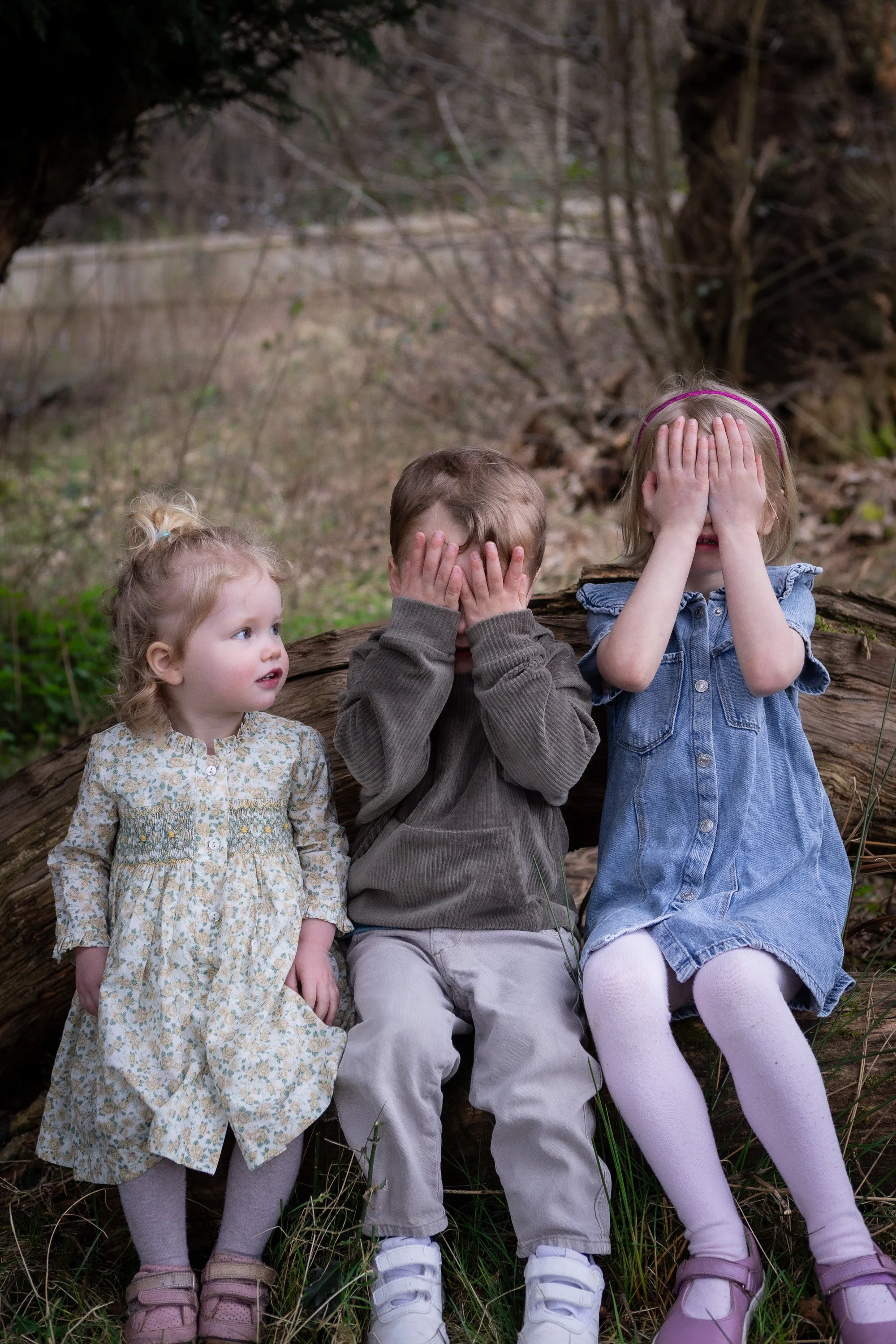 Three young children sitting outdoors behind a fallen log in a wooded area. The girl on the left is wearing a floral dress and socks with sandals, looking to the side. The boy in the middle is wearing a brown jacket and white sneakers, covering his f