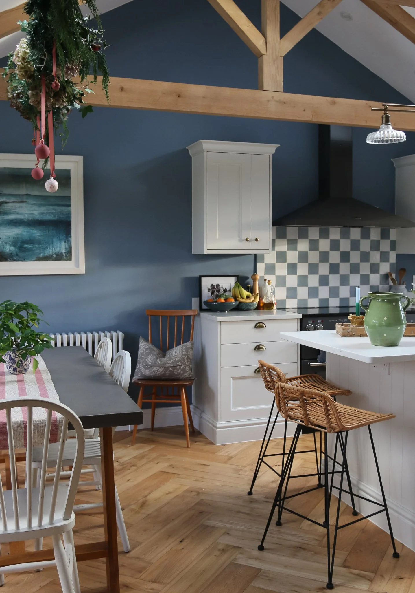 A cozy kitchen and dining area with wooden flooring, white cabinets, a blue painted wall, and a wooden beam ceiling. There is a black dining table with white chairs, a kitchen island with rattan and black chairs, a basket of bananas, and a green pitc