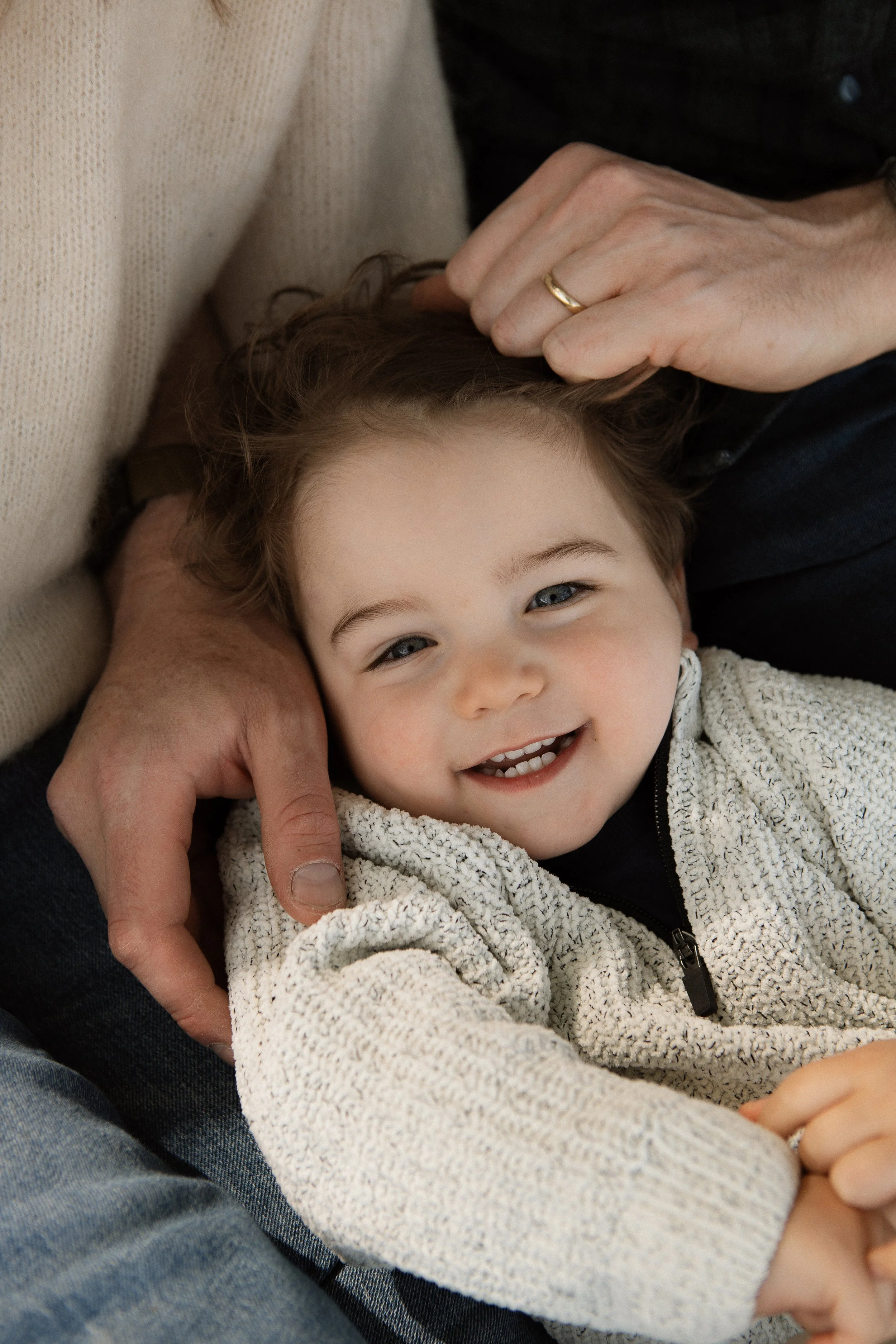 Close-up of a young boy with blue eyes smiling, being cuddled by an adult with a hand gently stroking his head, wearing a beige sweater and a white fleece jacket.