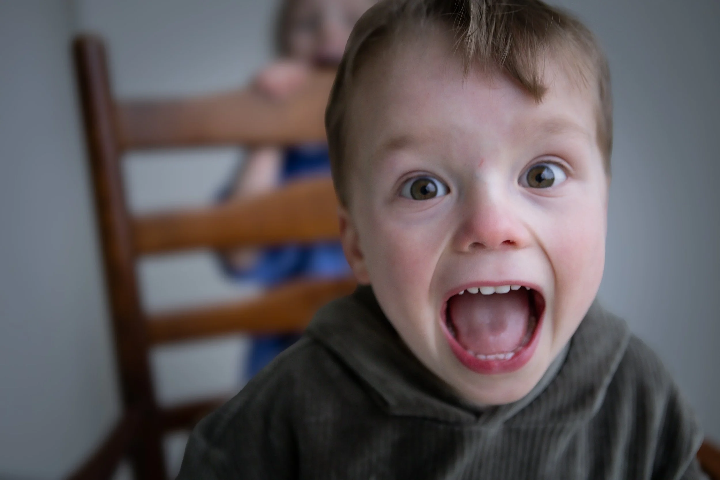 Close-up of a young boy with brown hair and hazel eyes, screaming or shouting with his mouth wide open. A blurred wooden chair can be seen in the background.