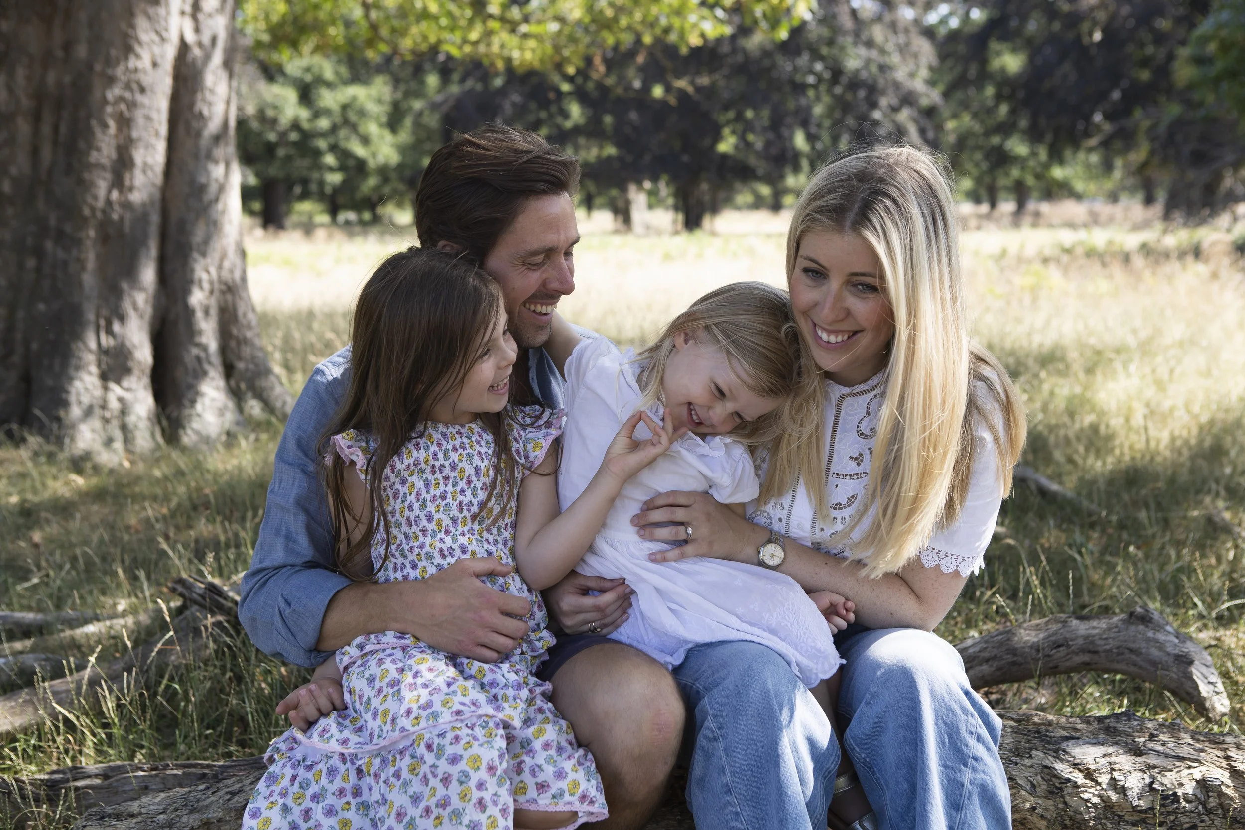 A family of five sitting on a log in a park, smiling and hugging each other, surrounded by trees and grass.