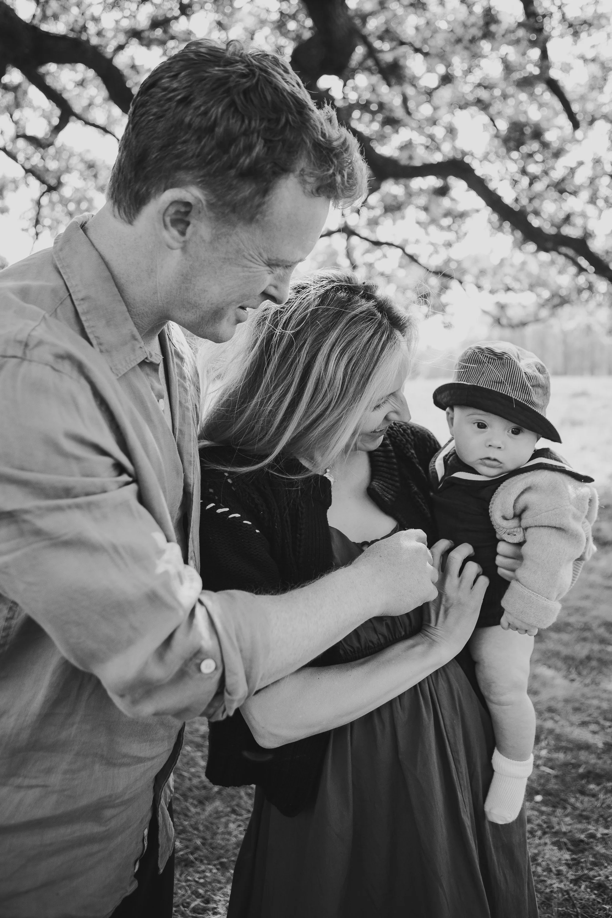 Three people, an adult man, a woman, and a young child, outside in a park or nature area, with trees in the background. They are looking at each other and smiling, and the woman is holding the child.