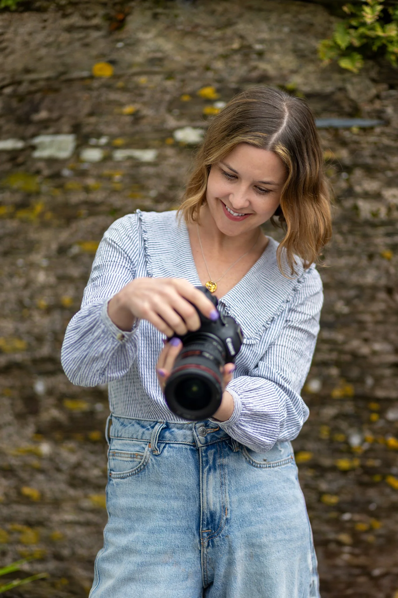 A young woman with shoulder-length wavy blonde hair wearing a gray and white striped long-sleeve shirt and high-waisted blue jeans holding a camera and smiling, outdoors in front of a blurred background with brown dirt and fallen yellow leaves.