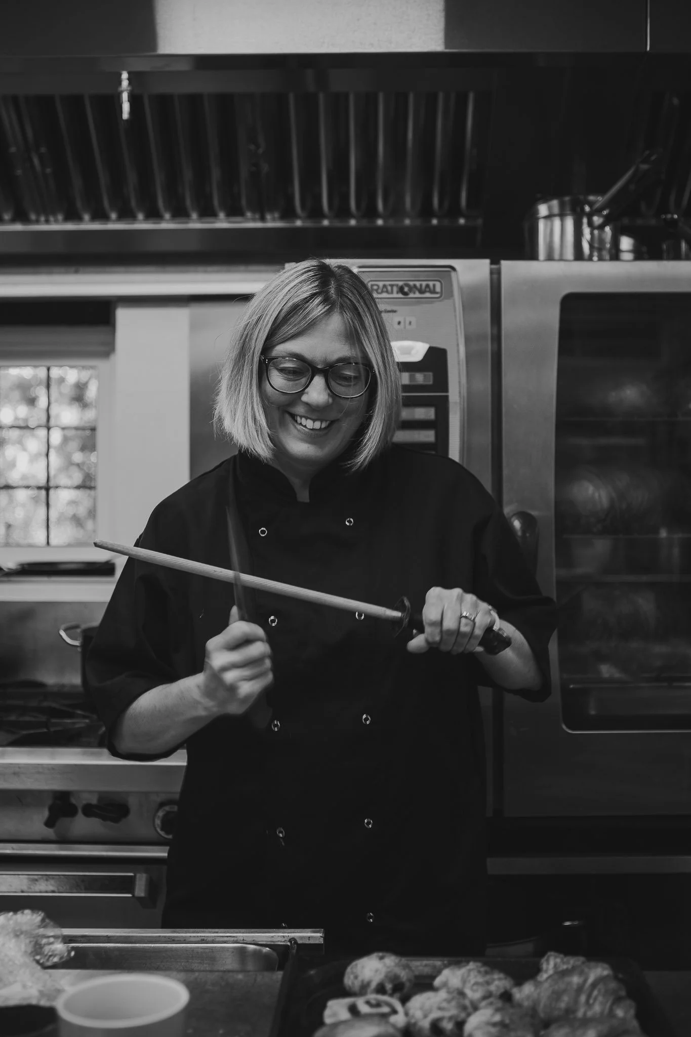 A woman in a black chef's coat with glasses, smiling and holding a cooking utensil in a commercial kitchen.