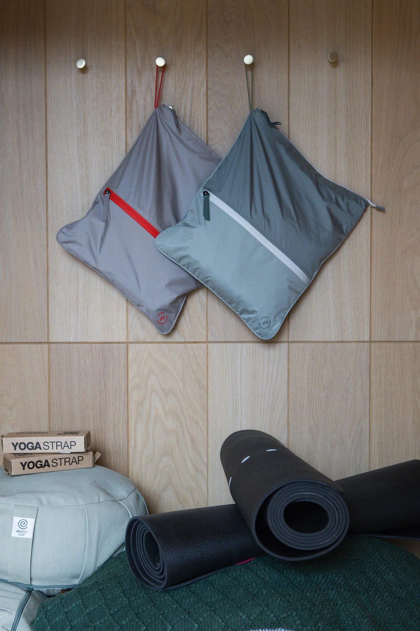 Two yoga mats, one black and one dark blue, rolled up and placed on a green carpet, with a grey yoga bag nearby. In the background, there are two packages labeled "YOGA STRAP" leaning against the wall.
